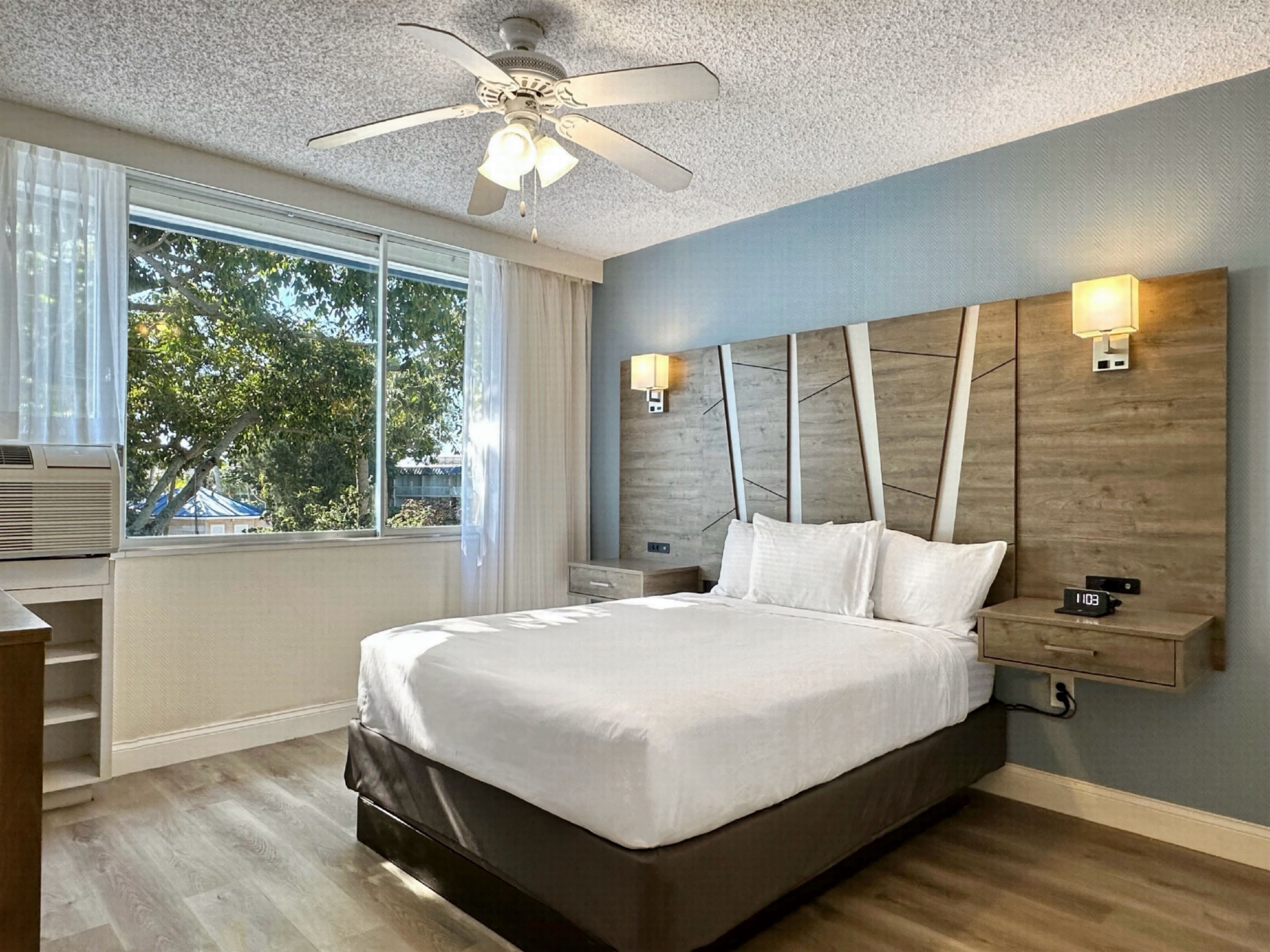 Un dormitorio moderno con una cama grande, un ventilador de techo, una mesita de noche con lámpara y una ventana con cortinas.