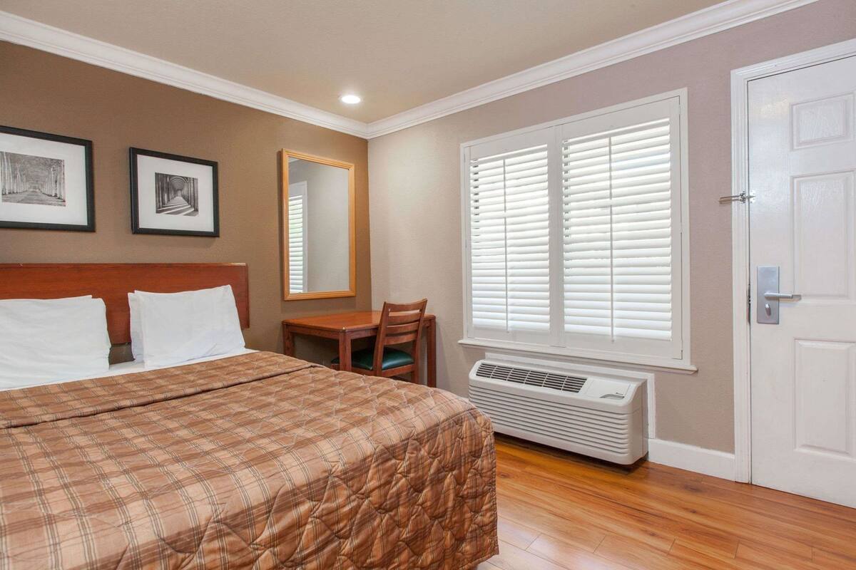 Une chambre d’hôtel équipée d’un lit, d’un bureau, d’une chaise, d’un miroir et d’une fenêtre avec des stores.