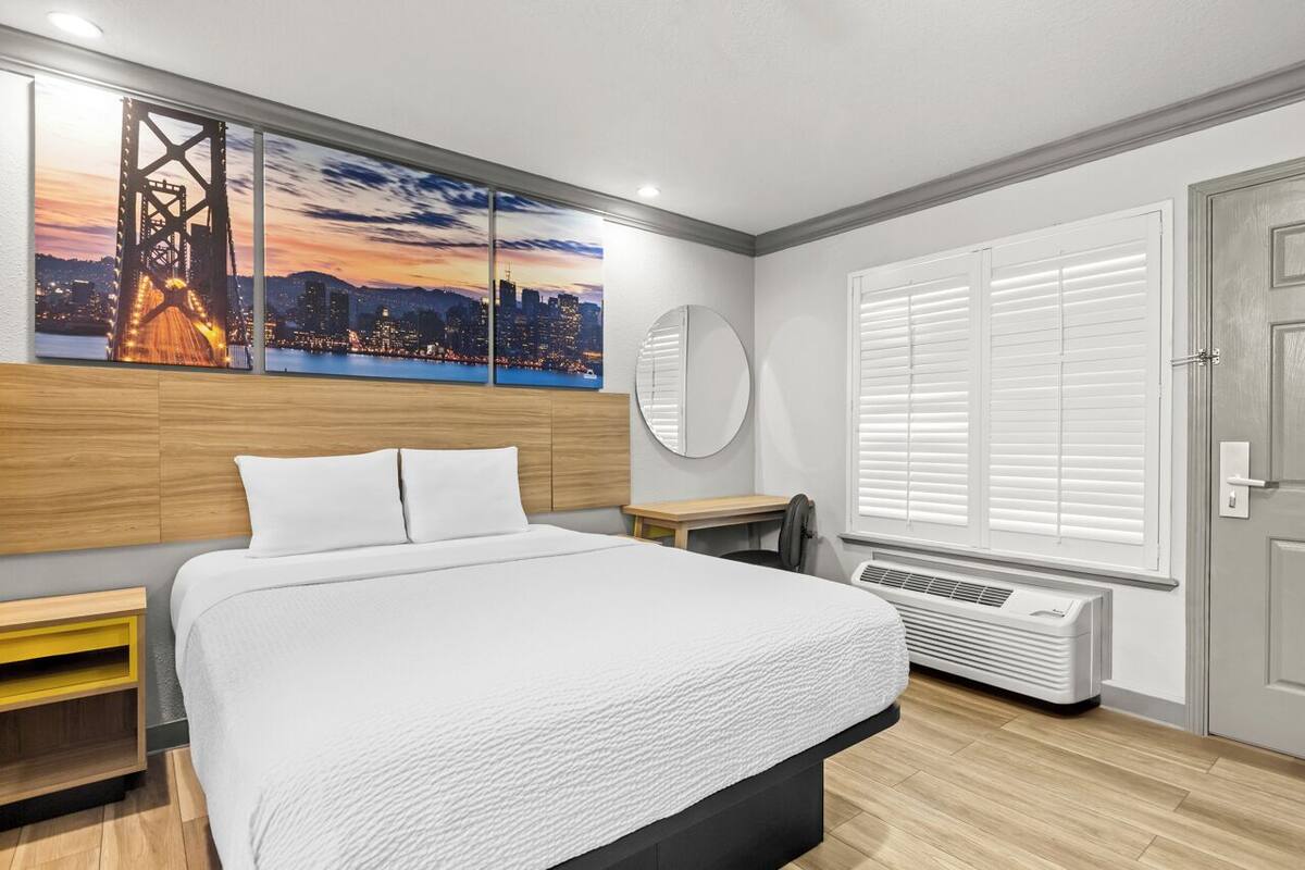 Une chambre d’hôtel moderne avec un grand lit, un tableau représentant un paysage urbain, un miroir rond, un radiateur et une fenêtre avec des stores.