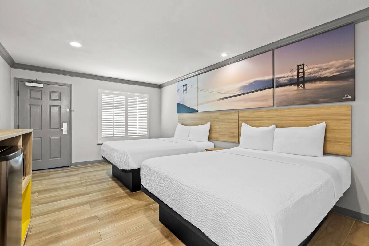 Une chambre d’hôtel avec deux lits, un plancher en bois, une porte grise et un grand tableau mural.