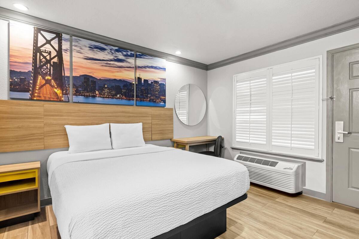 Une chambre d’hôtel moderne avec un grand lit, une vue sur la ville, un miroir rond et une fenêtre avec des stores.