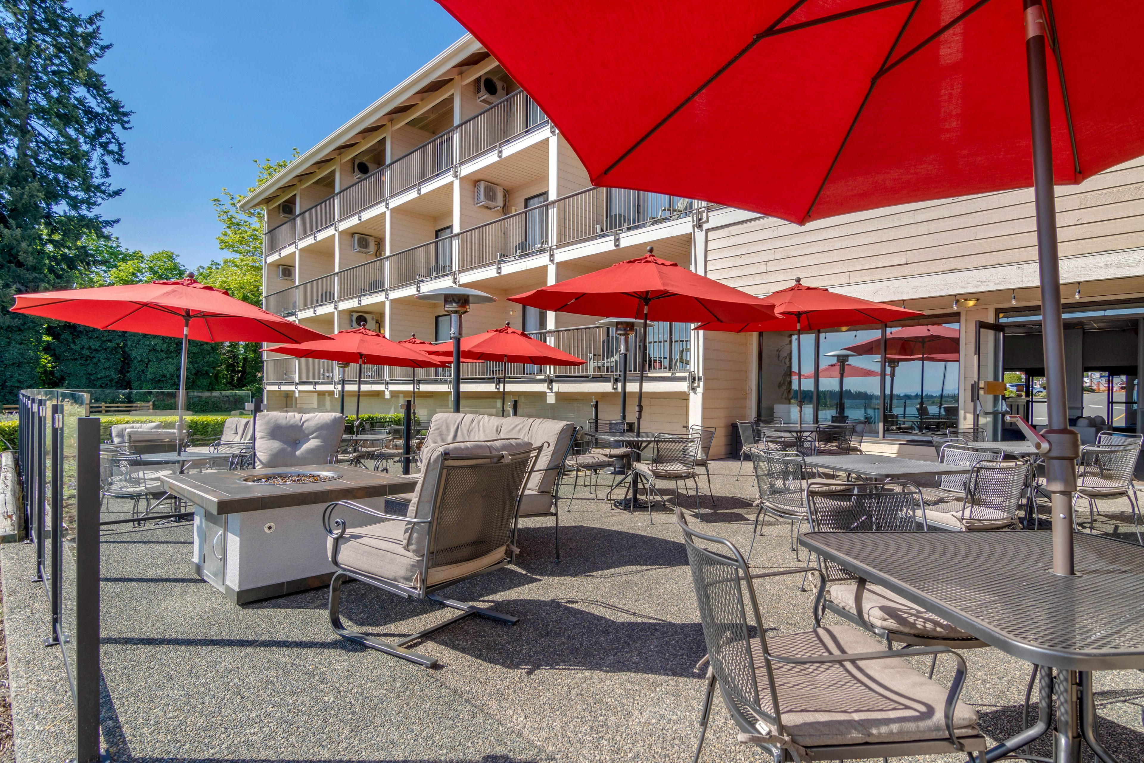 An outdoor patio area with multiple tables and chairs, red umbrellas, and a building with balconies in the background.