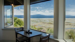 A room with a large window offering a view of a beach and mountains. There is a table with chairs and a rolled-up mat on it.