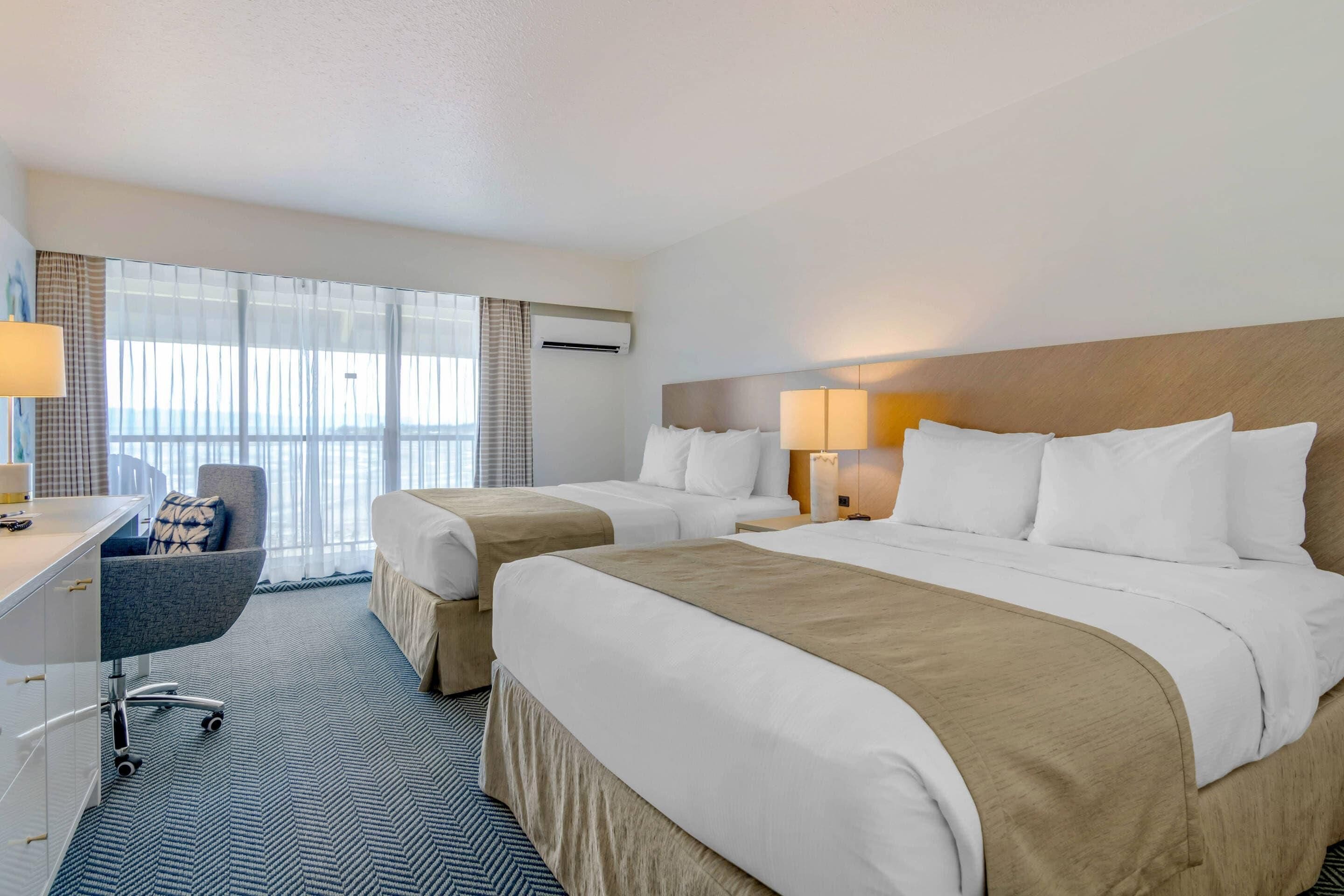 Habitación de hotel con dos camas, un escritorio y una silla. Cuenta con un ventanal con cortinas, una lámpara y un equipo de aire acondicionado.
