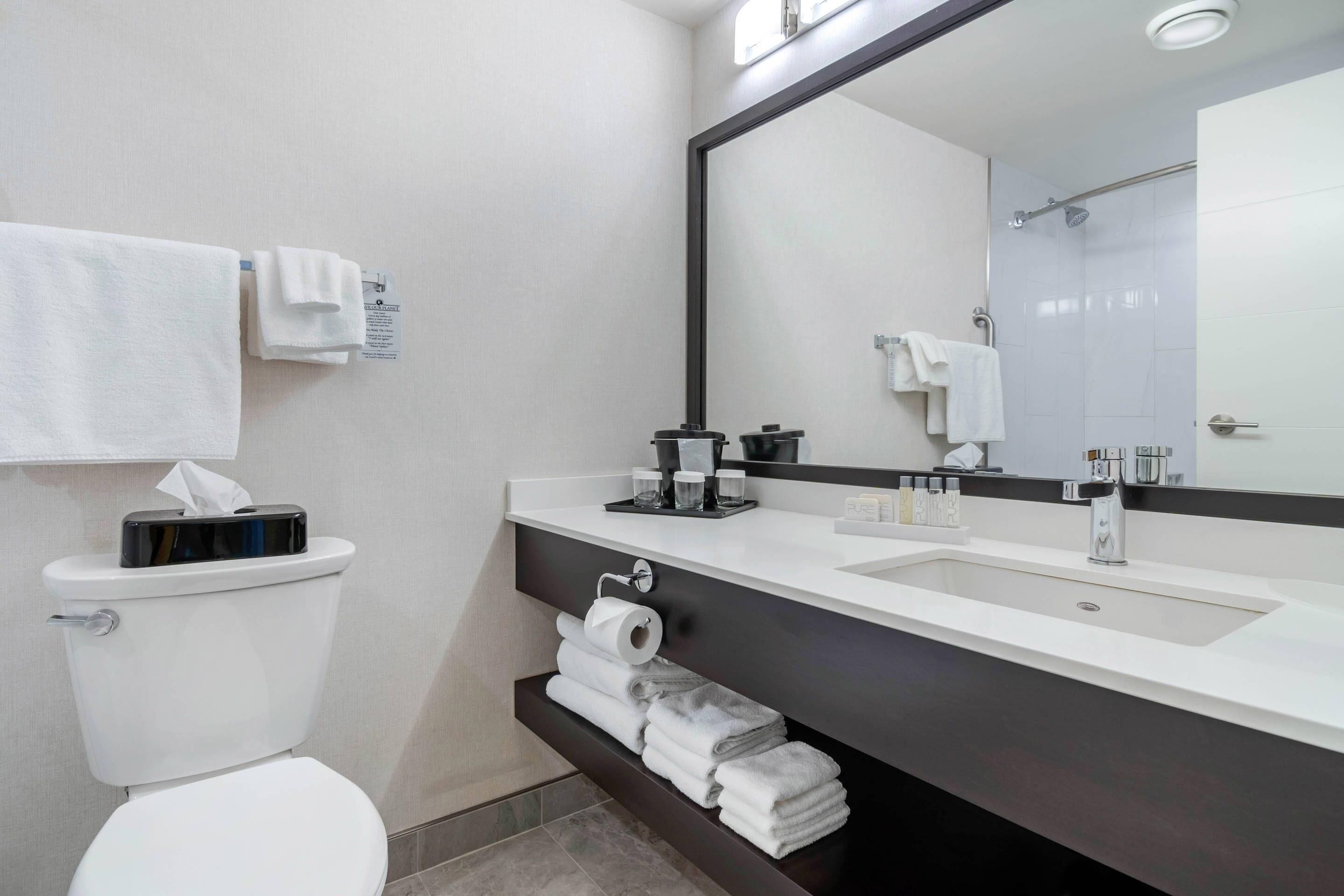 Une salle de bain moderne avec des toilettes blanches, un meuble-lavabo noir, un grand miroir et des serviettes soigneusement pliées.