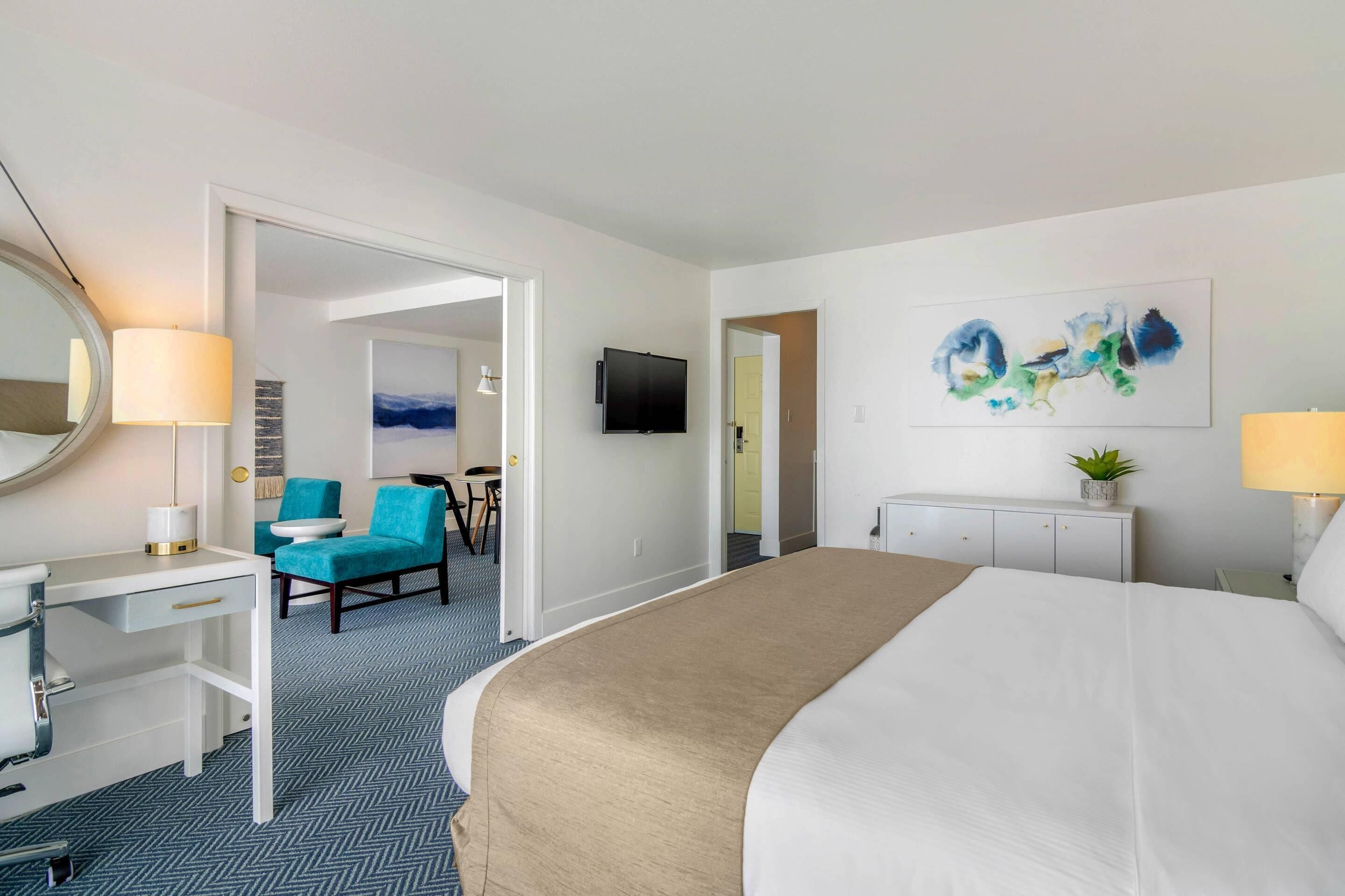A hotel room with a bed, a desk, a chair, a television, and a painting on the wall.