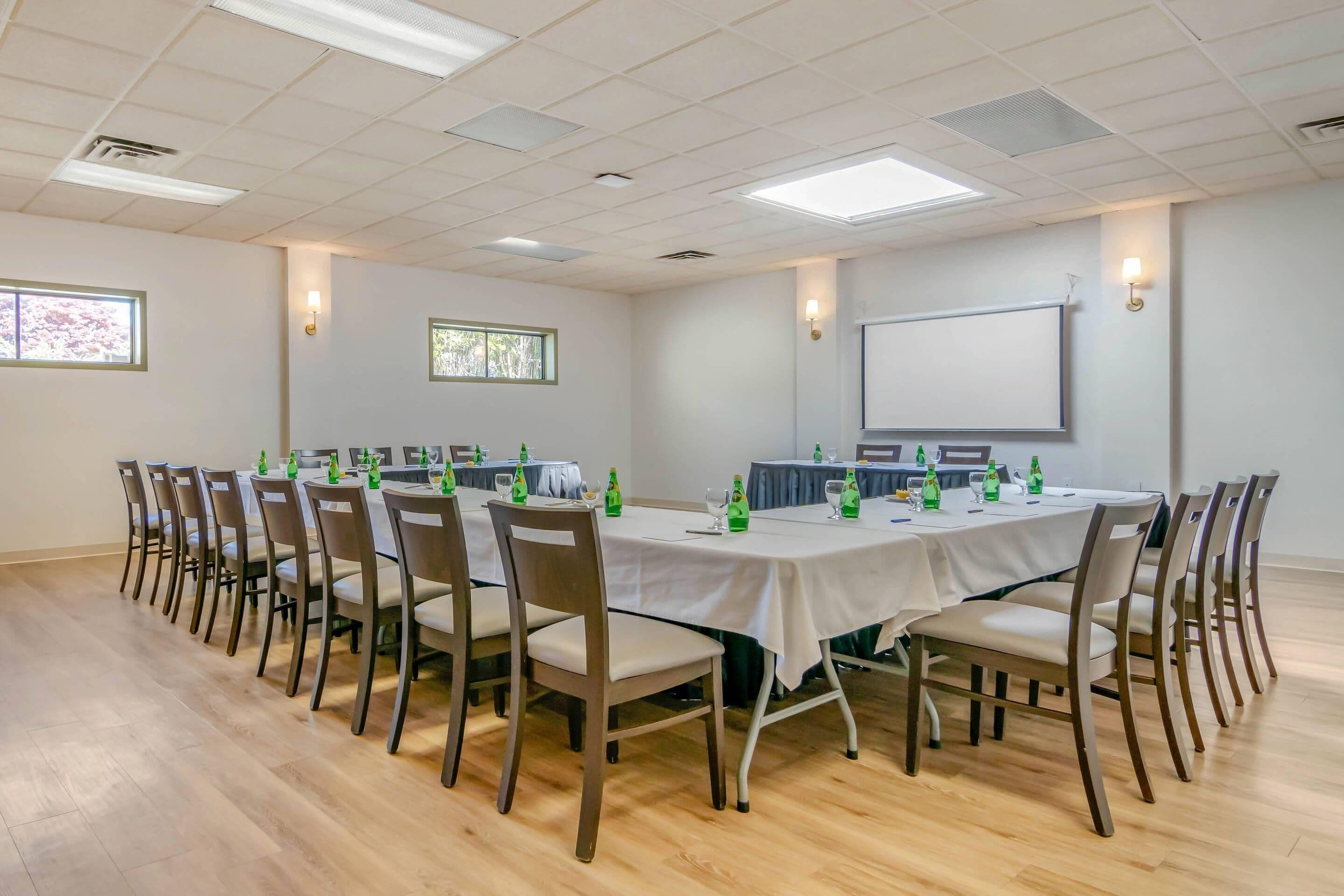 A conference room with a long table, chairs, a projector screen, and two windows.
