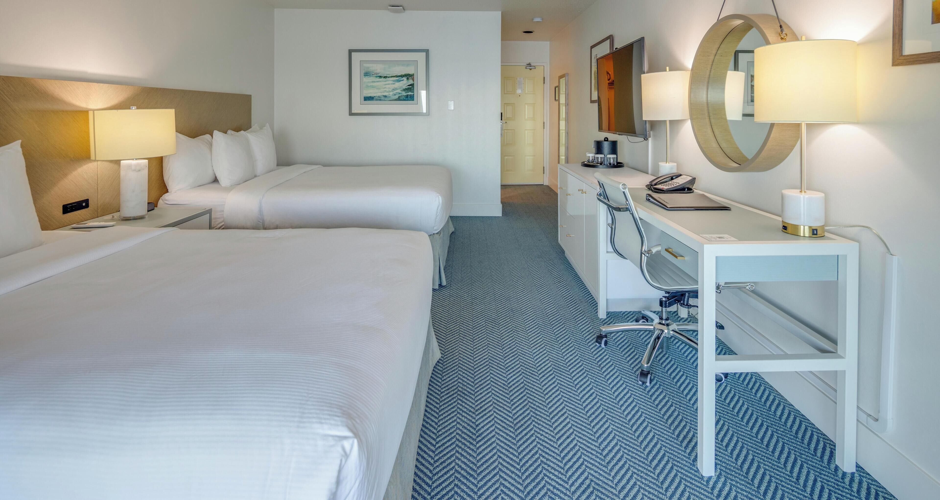 A hotel room with two beds, a desk with a chair, a desk lamp, a telephone, and a mirror.