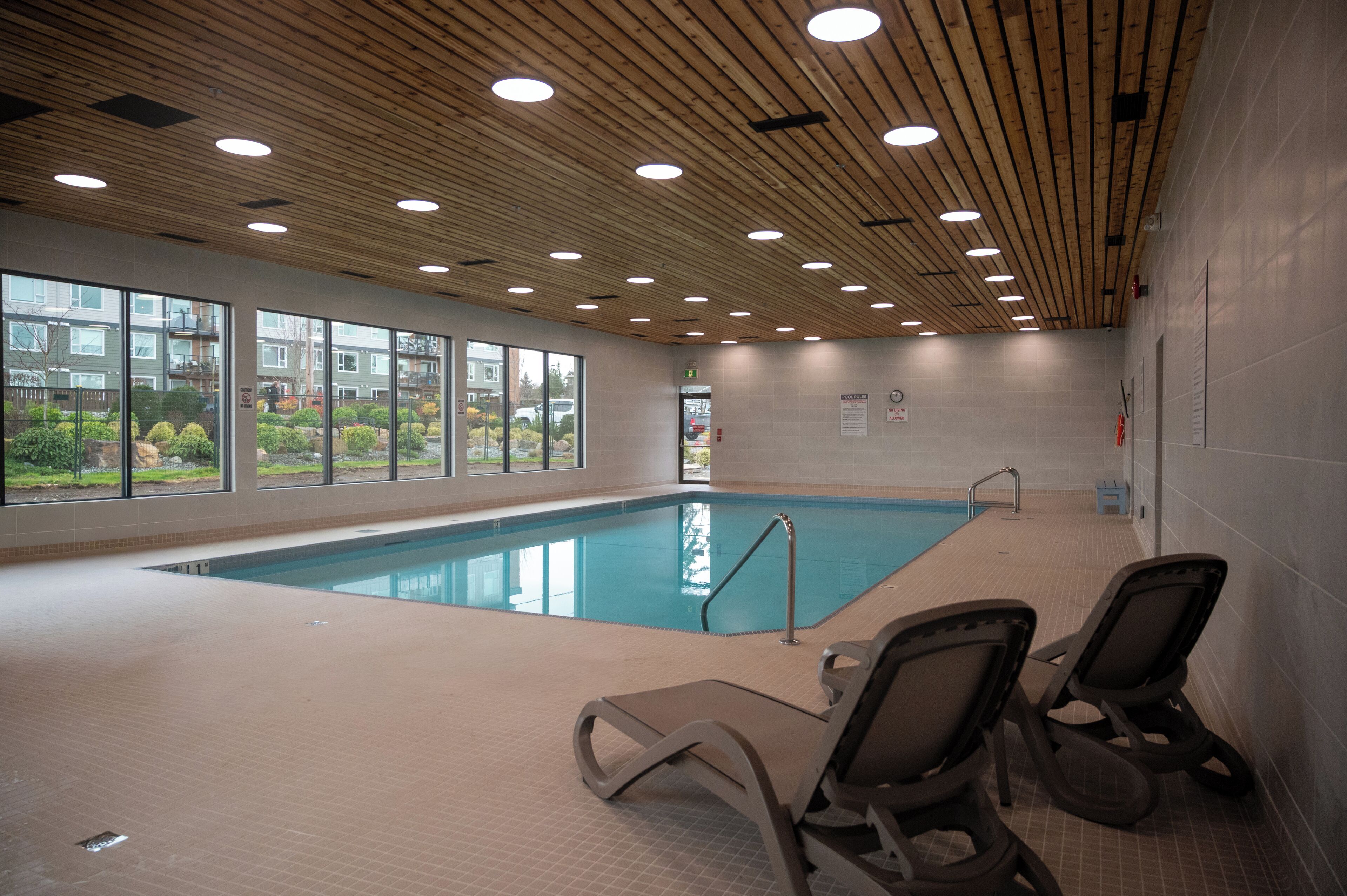 An indoor swimming pool with wooden ceiling, clear windows, and lounge chairs.