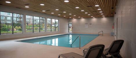 An indoor swimming pool with wooden ceiling, clear windows, and lounge chairs.