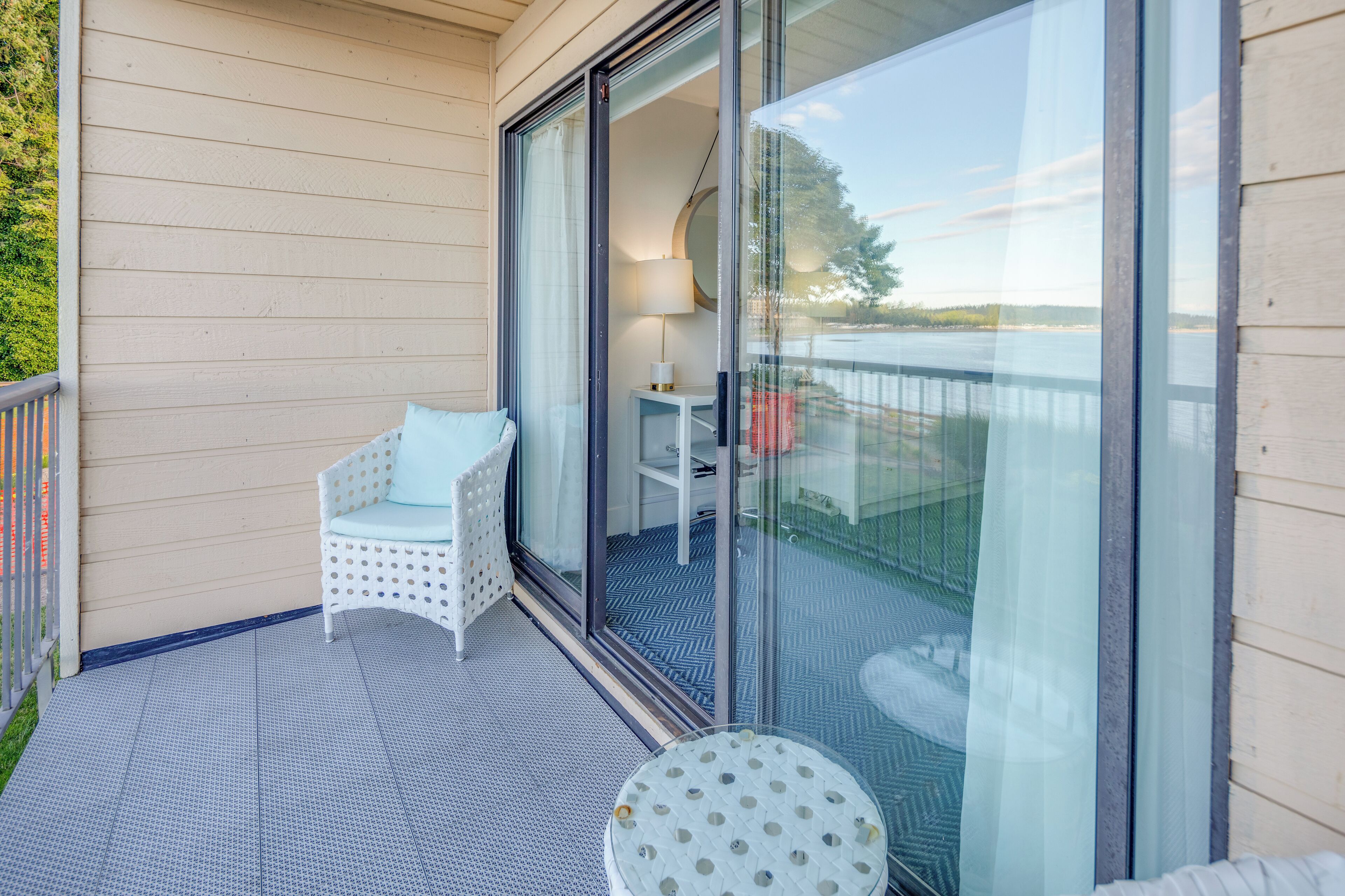 A balcony with a white chair and a small table, a glass door leading to a room, and a view of a body of water.