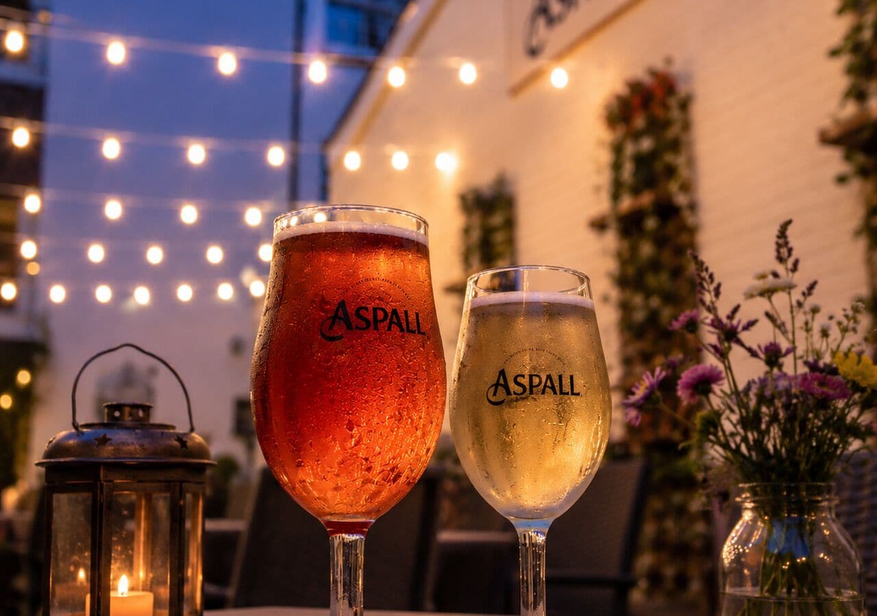 Due bicchieri di birra Aspall su un tavolo, con una lanterna e un vaso di fiori sullo sfondo.