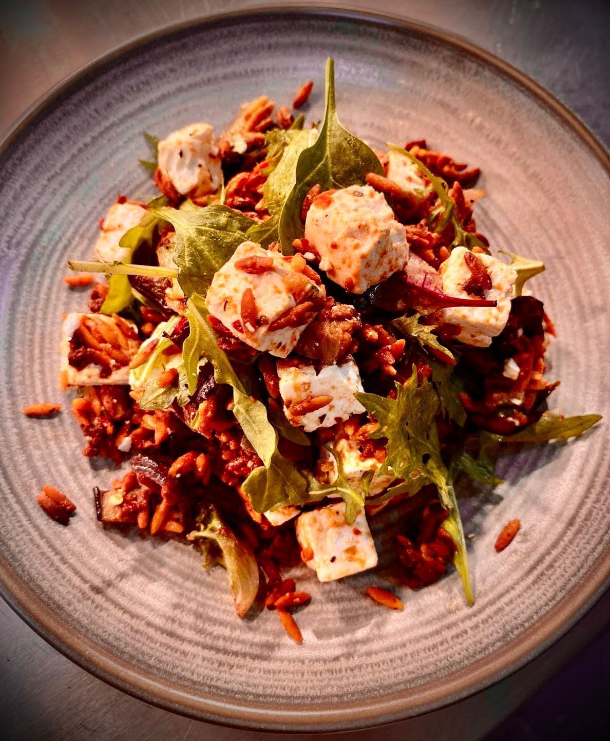 Une assiette remplie de divers aliments, notamment des légumes verts, des légumes rouges et des cubes d’une substance blanche, qui pourrait être du fromage ou du tofu, le tout présenté sur une assiette en céramique à texture particulière.