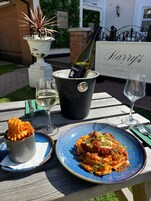 Mesa al aire libre con botella de vino, copas, un plato de pasta y un bol de papas fritas.