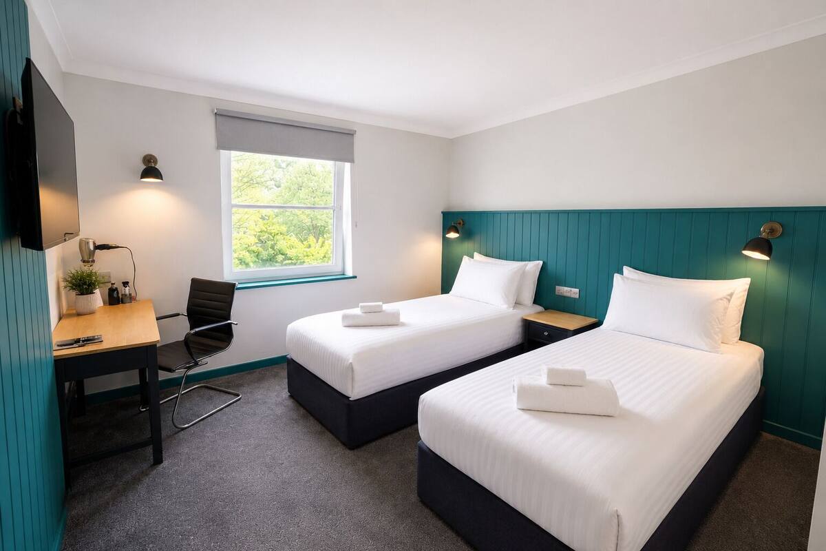 Ein Hotelzimmer mit zwei Betten, einem Schreibtisch, einem Stuhl, einem Fenster und einem Fernseher.