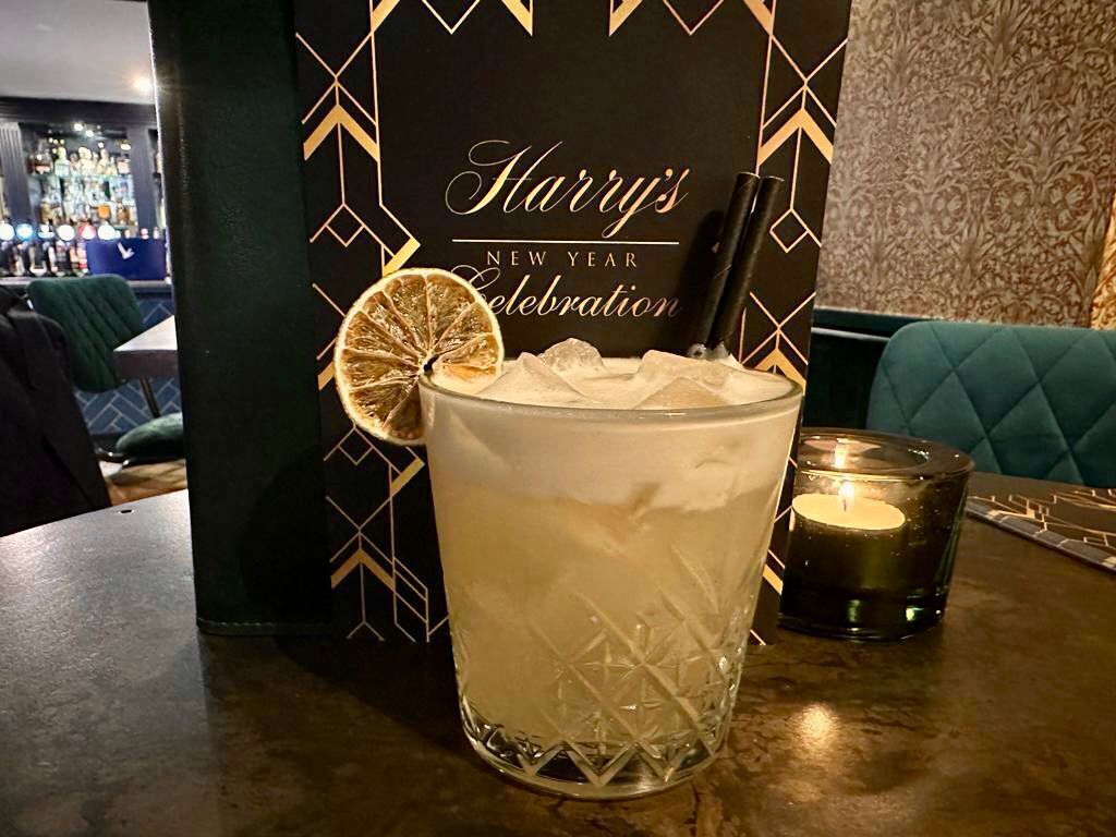A glass of a clear beverage with a lemon slice garnish, ice cubes, and a straw, placed on a table with a dark patterned backdrop and a candle.