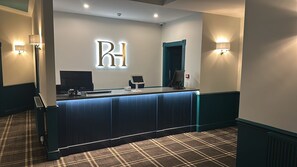Bancada de recepção de hotel com um grande letreiro iluminado 'RH', um computador, um telefone e uma pequena área de estar.