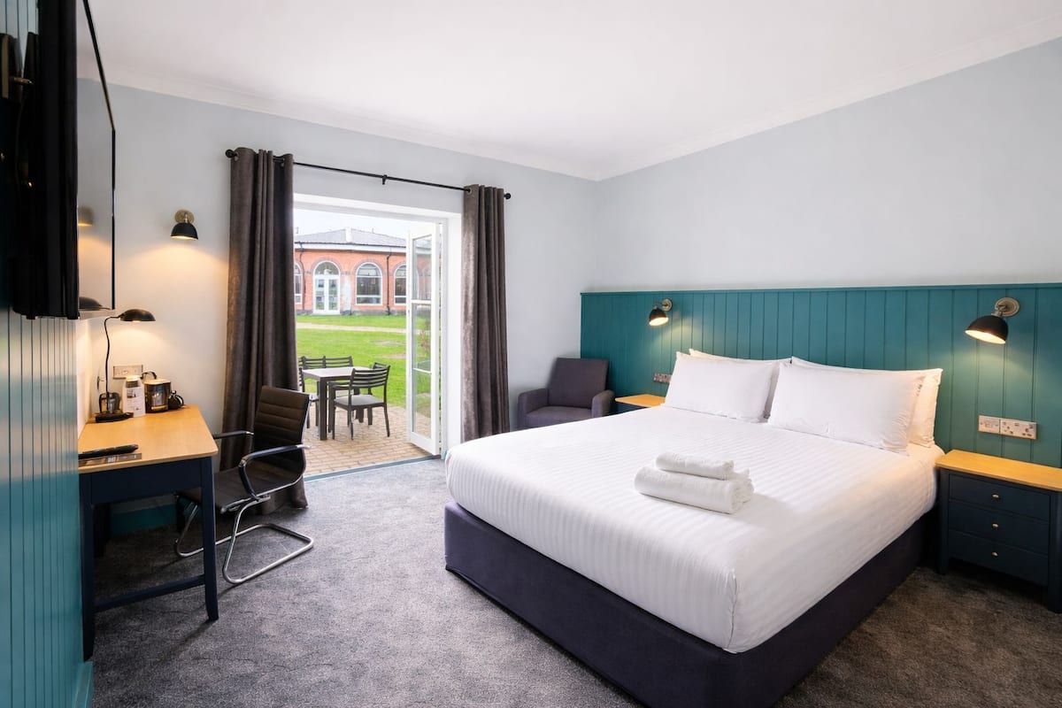 Ein Hotelzimmer mit Bett, Schreibtisch, Stuhl und Blick auf ein Gebäude durch eine Glastür.