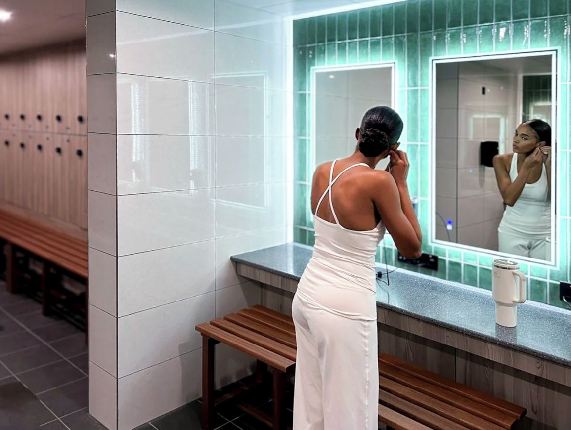 A woman in a white dress standing in front of a mirror in a bathroom with a bench and lockers in the background.