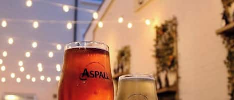 Dos vasos de cerveza Asphall, uno roja y otro blanca, sobre una mesa con luces de cuerda al fondo.