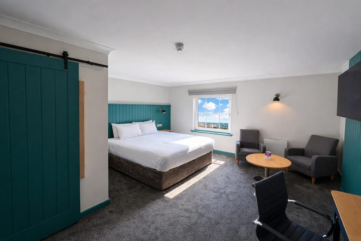 Ein Hotelzimmer mit einem Bett, zwei Sesseln, einem kleinen Tisch, einem Fenster mit Blick und Wänden in Türkis.
