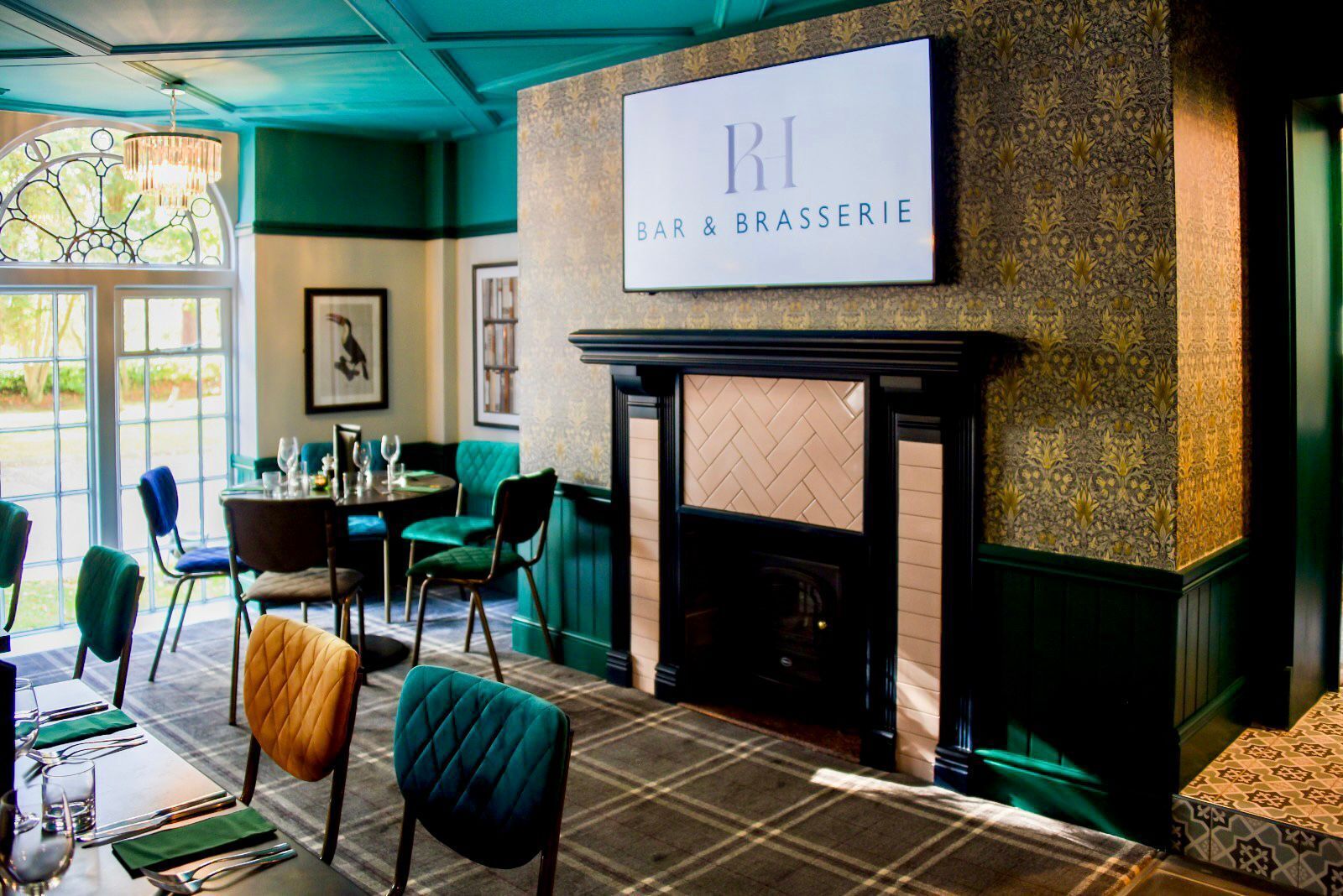 A bar and brasserie with a fireplace, green upholstered chairs, and a plaid carpet.