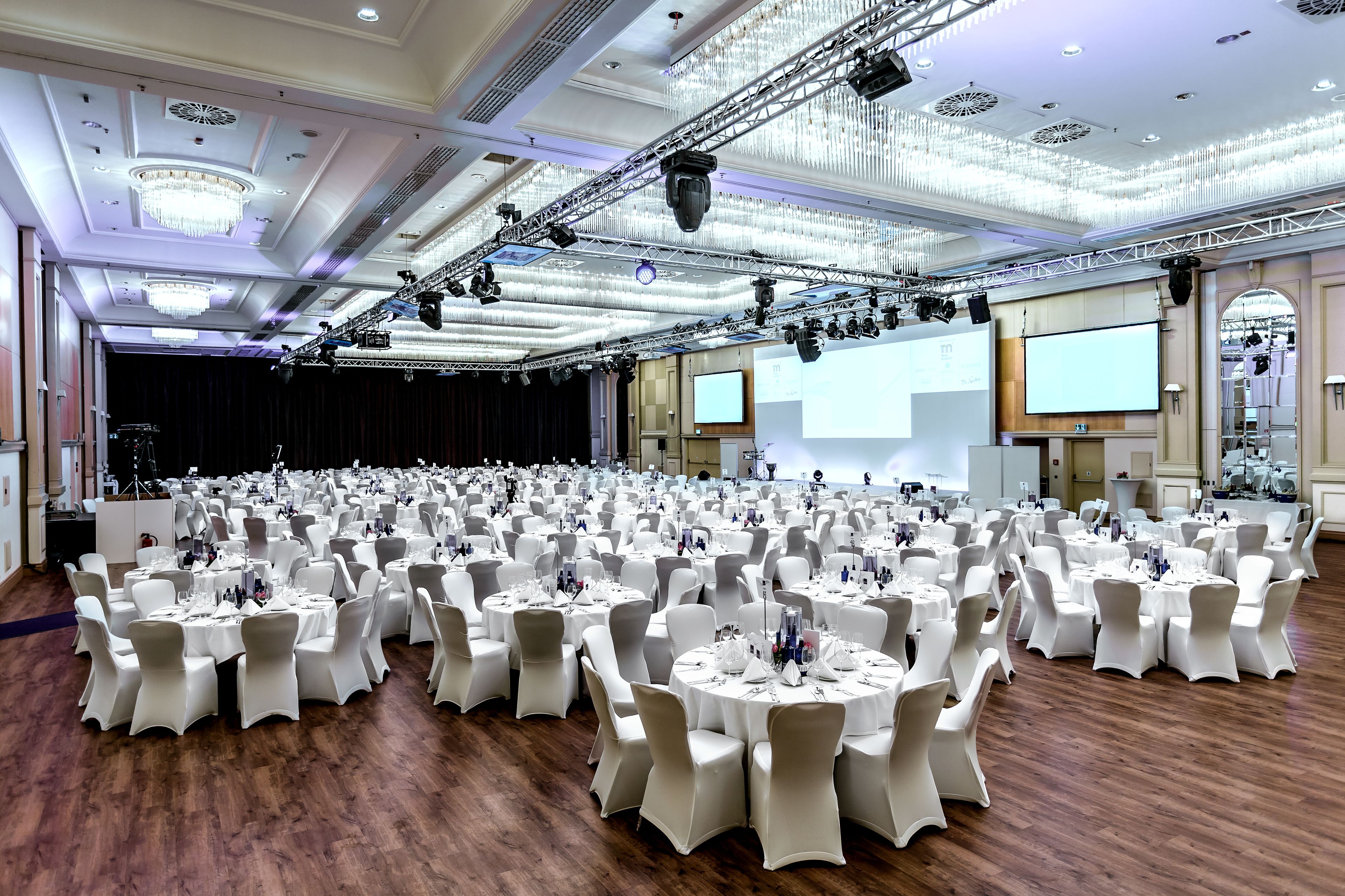Un amplio salón de banquetes con mesas redondas dispuestas para eventos, equipado con iluminación y pantallas de proyección.