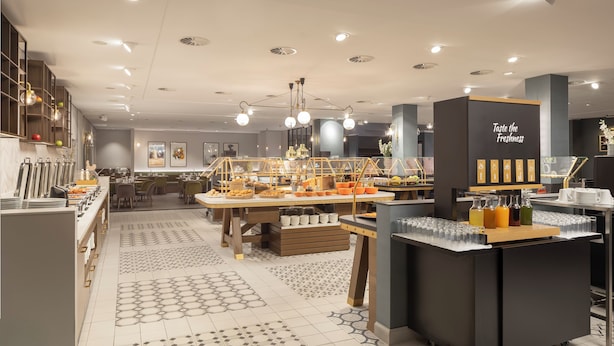 A modern cafe with a buffet setup, including a variety of food items and beverages, and a sign that reads "Taste the Freshness." - Hilton Düsseldorf (Düsseldorf)