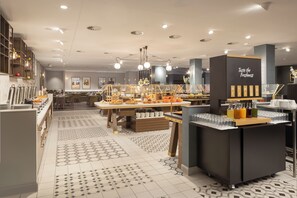 A modern cafe with a buffet setup, including a variety of food items and beverages, and a sign that reads "Taste the Freshness." - Hilton Düsseldorf (Düsseldorf)