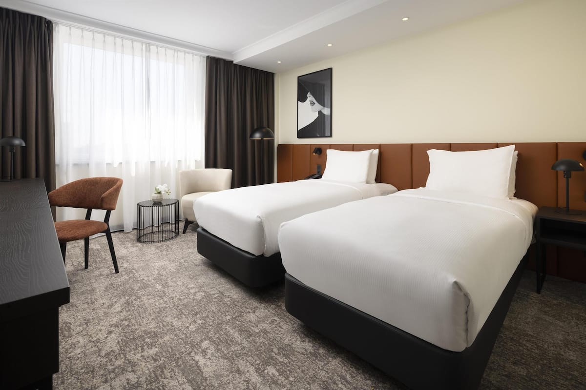 Een hotelkamer met twee bedden, een bureau, een stoel, een tafeltje en een groot raam met gordijnen.