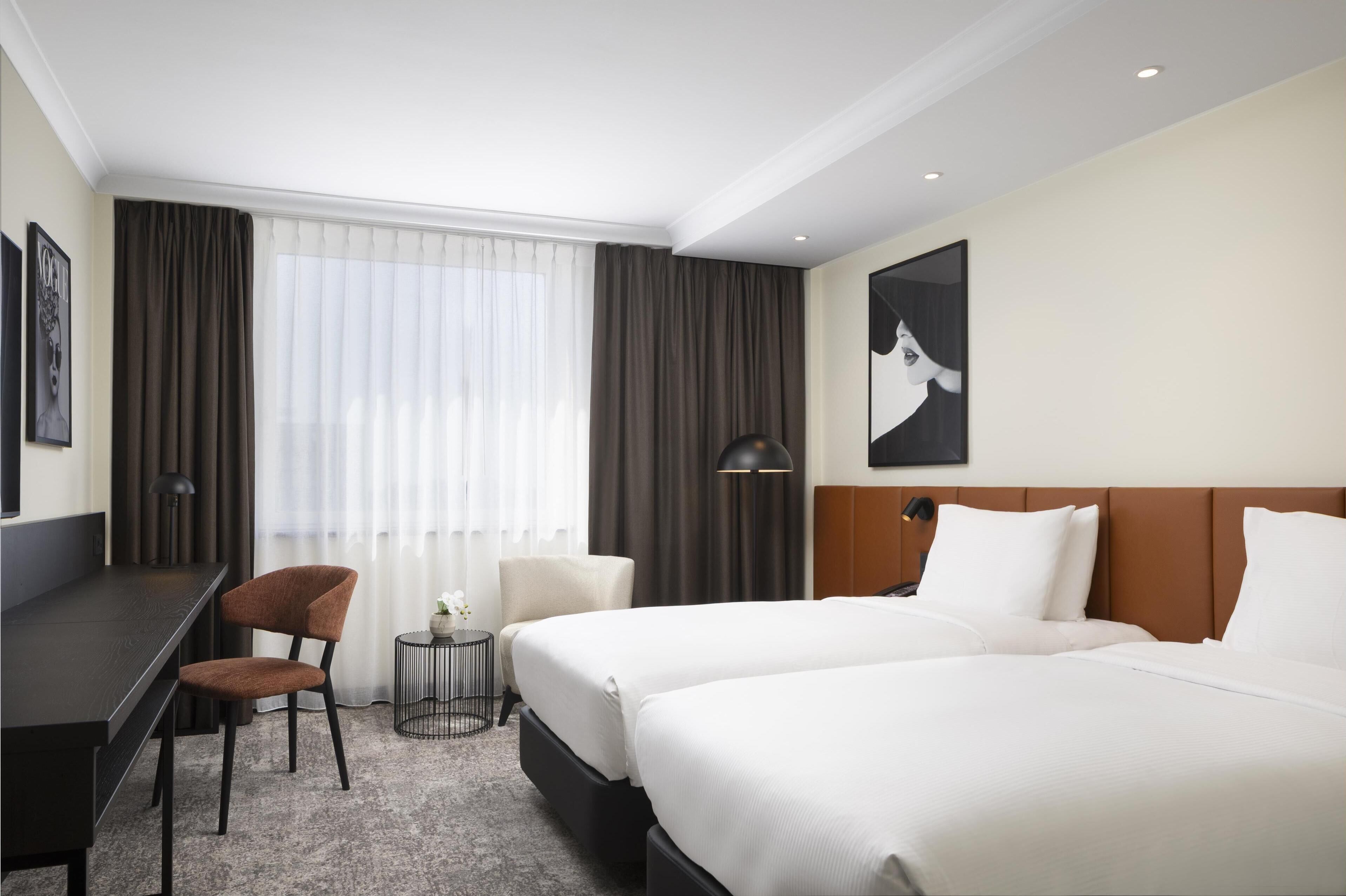 Een hotelkamer met twee bedden, een bureau, een stoel, een bijzettafeltje met een lamp en een groot raam met gordijnen.