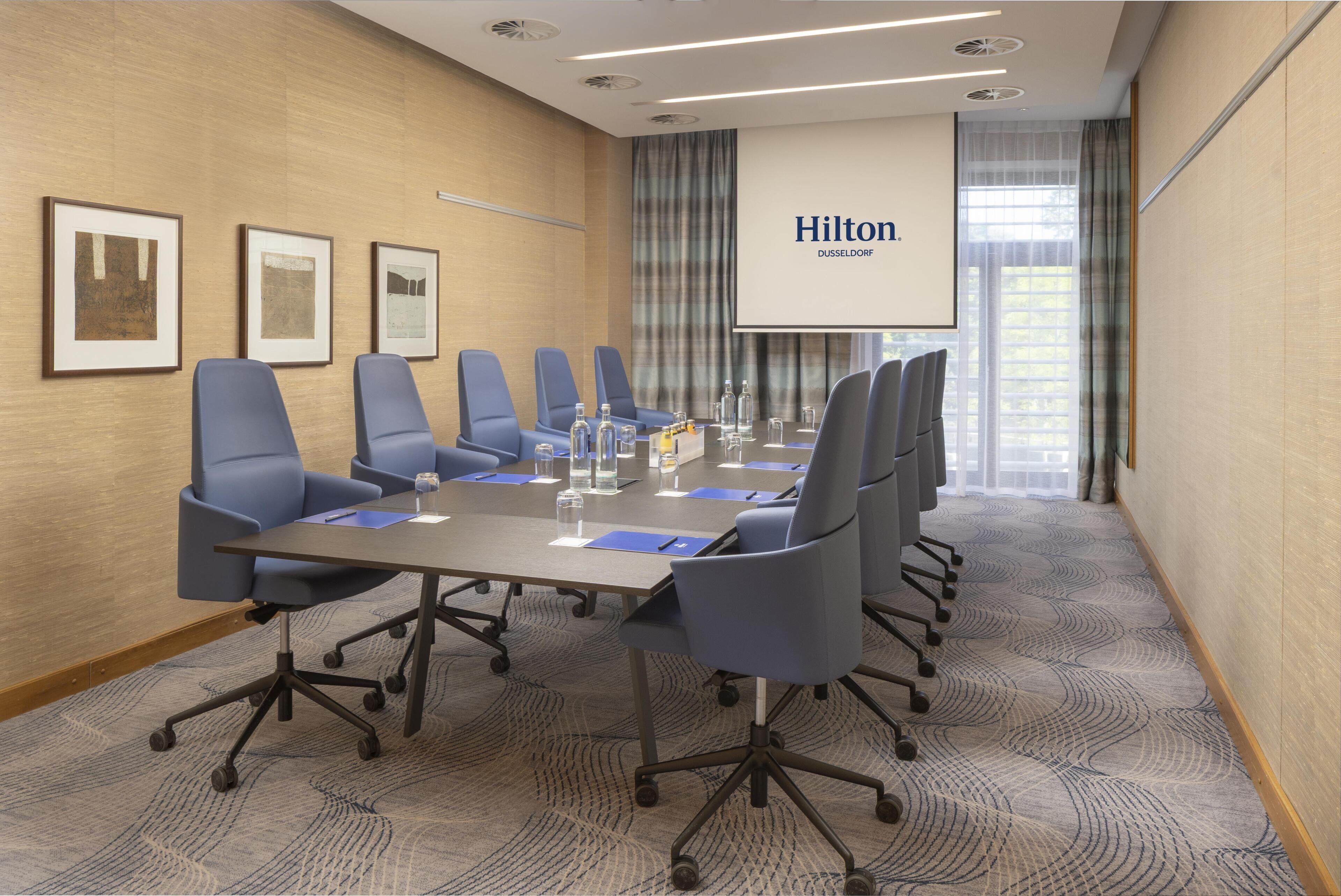 A conference room with a long table, six chairs, and a projector screen displaying 'Hilton DUSSELDORF'.