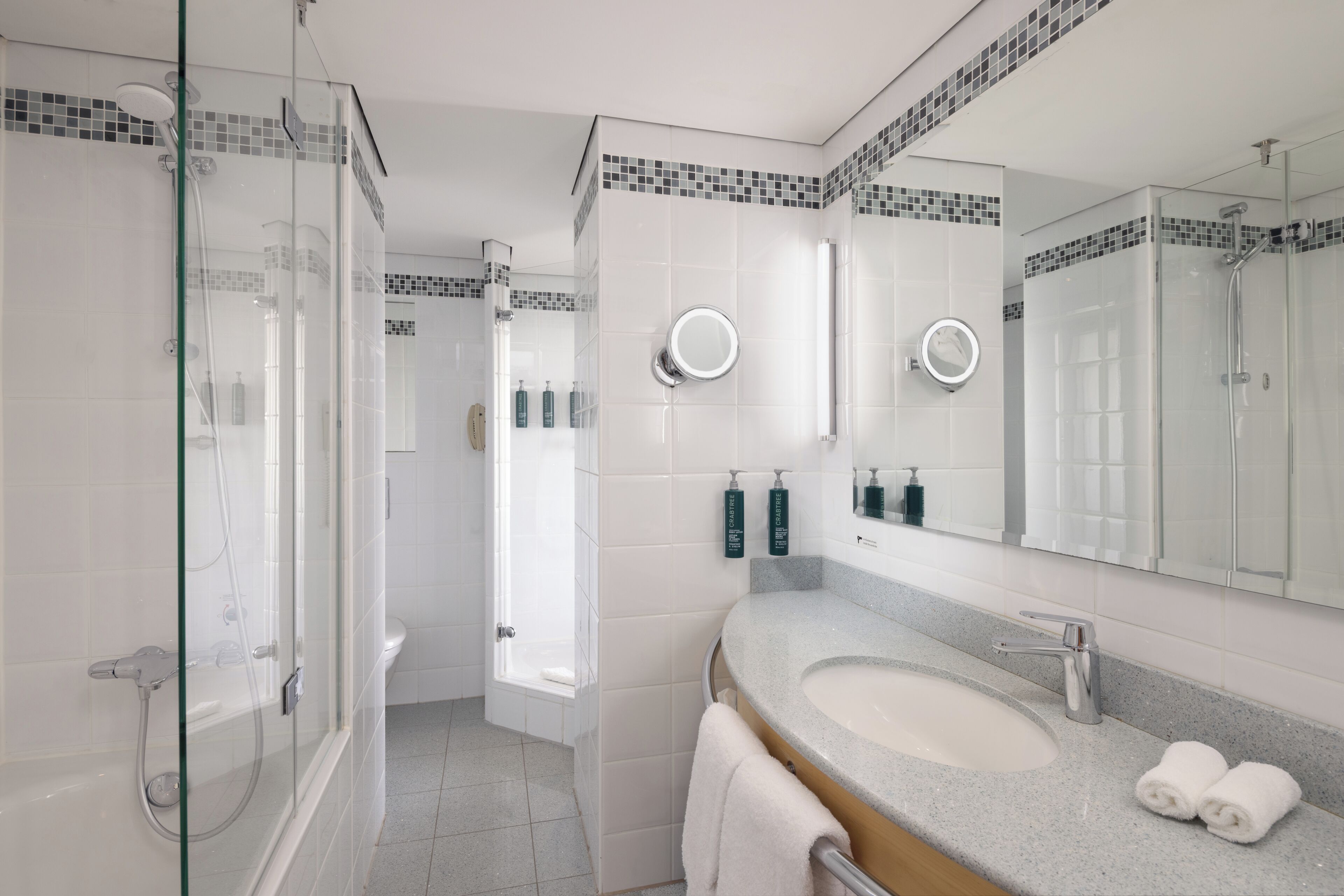 Un baño moderno con ducha de vidrio, lavamanos con tocador y espejo con luz redonda.