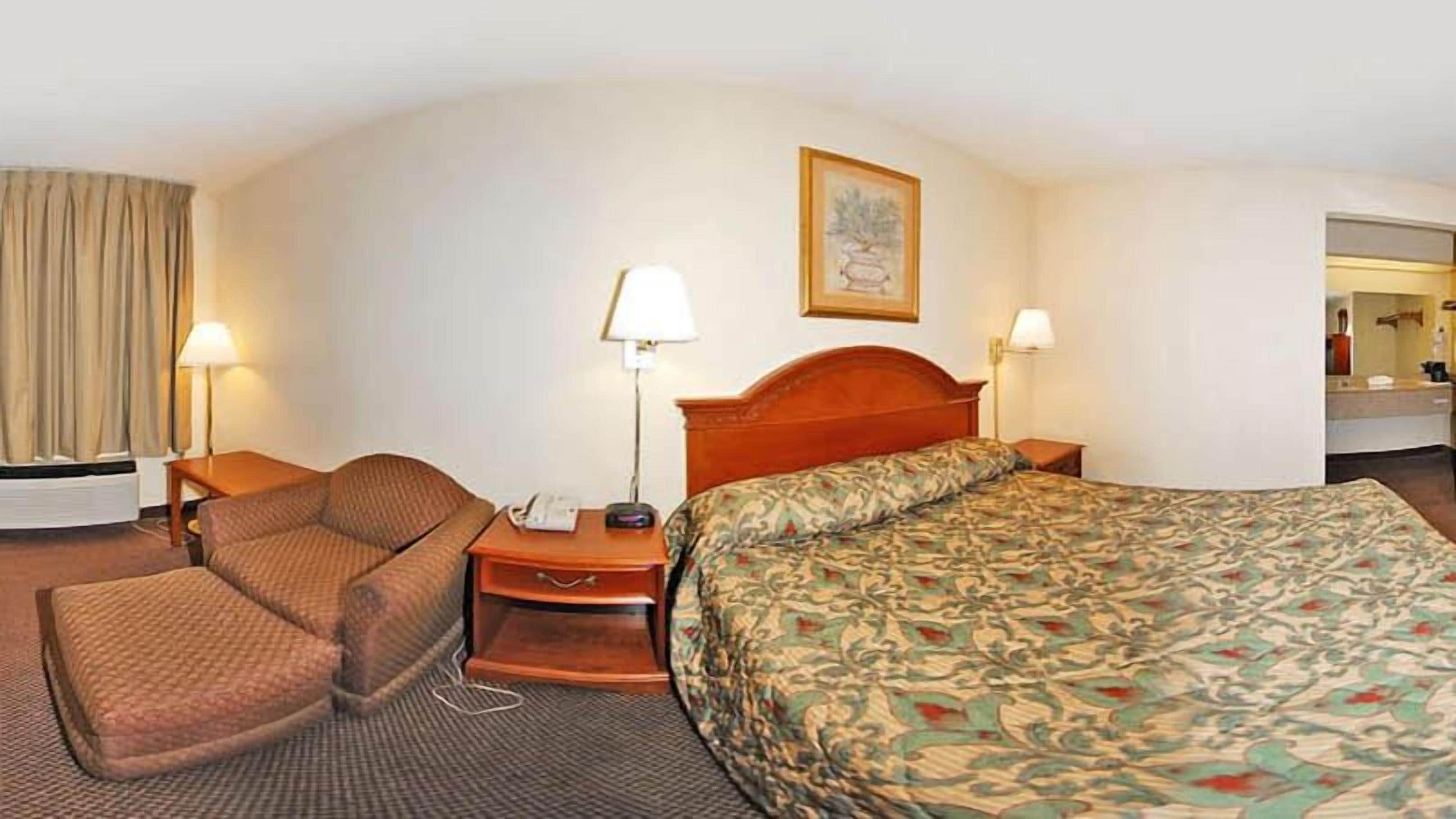 A hotel room with a bed, a chair, a desk with a lamp, and a painting on the wall.