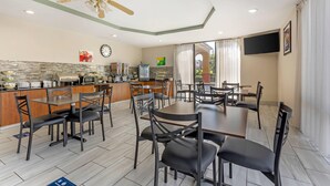 A spacious dining area with a kitchen in the background, featuring a modern design with a ceiling fan, a clock, and a flat-screen TV mounted on the wall.