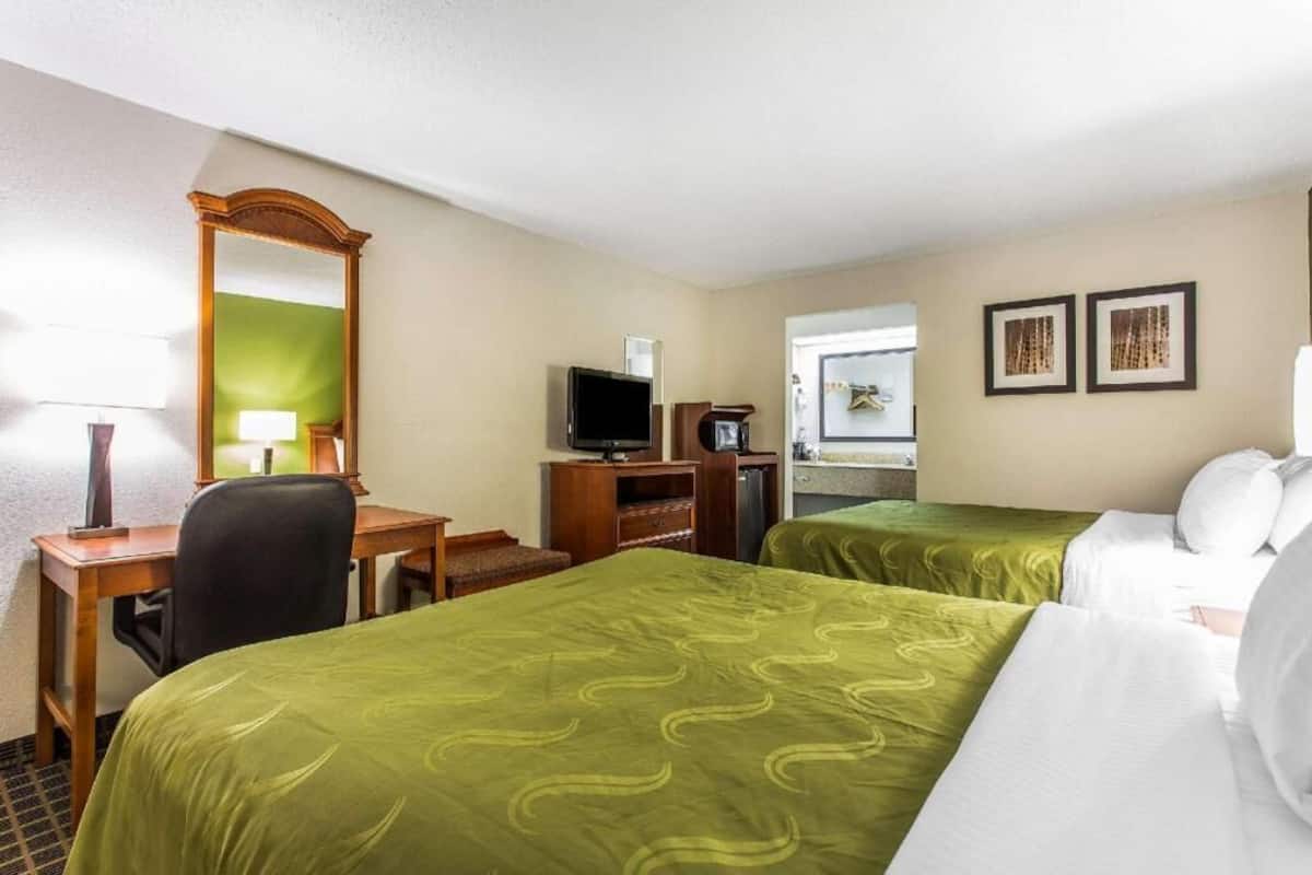 A hotel room with two beds, a desk, a television, a mirror, and two framed pictures on the wall.