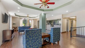 A spacious living area with a sofa, two patterned armchairs, a wooden coffee table, and a ceiling fan. A flat-screen TV is mounted on the wall, and a kitchen area is visible in the background.