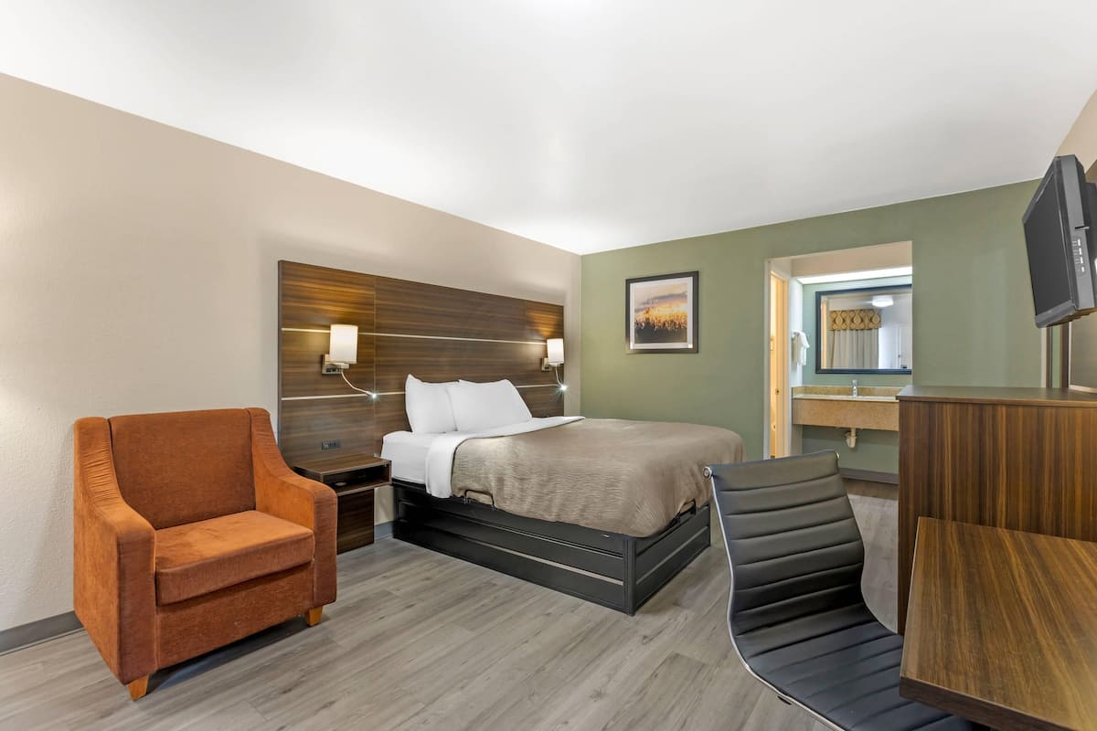 A hotel room with a bed, a brown armchair, a black chair, a wooden desk, and a television.