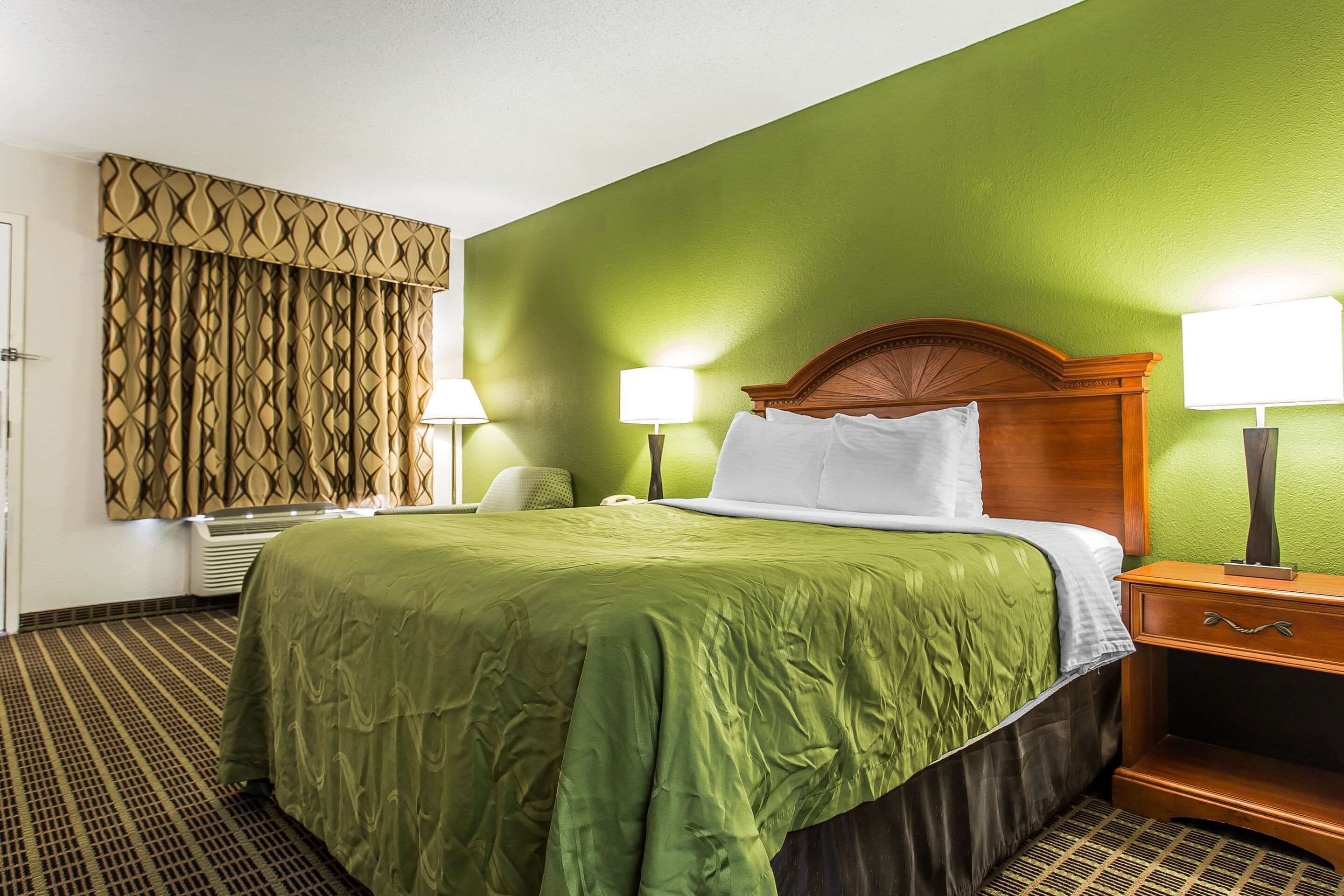 A hotel room with a green wall, a wooden headboard, a green bedspread, two bedside lamps, and a patterned curtain.