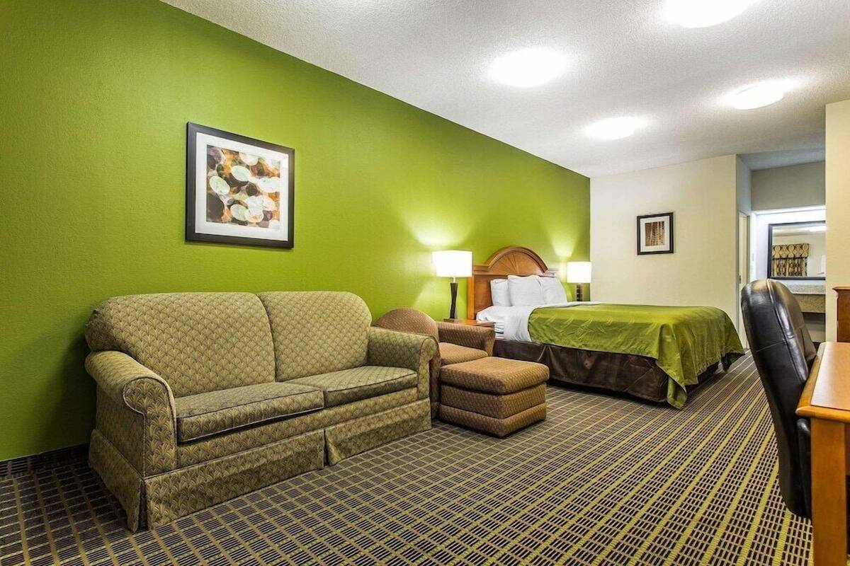 A hotel room with a green wall, a sofa, a chair, a bed with a green bedspread, a desk, and a lamp.