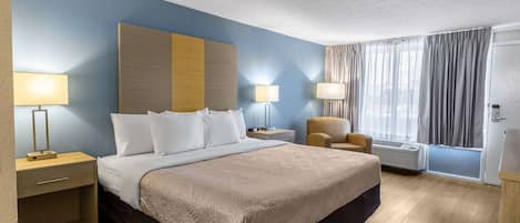A hotel room with a large bed, two bedside tables with lamps, a chair, an air conditioning unit, and a view of the outdoors through a window with curtains.