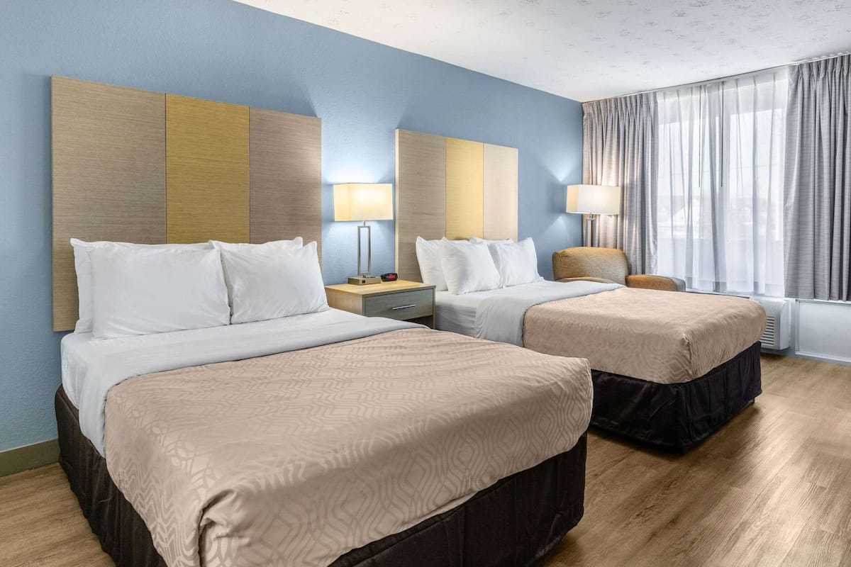 Ein Hotelzimmer mit zwei Betten, einem Nachttisch, einer Lampe, einem Sessel und einem Fenster mit Vorhängen.