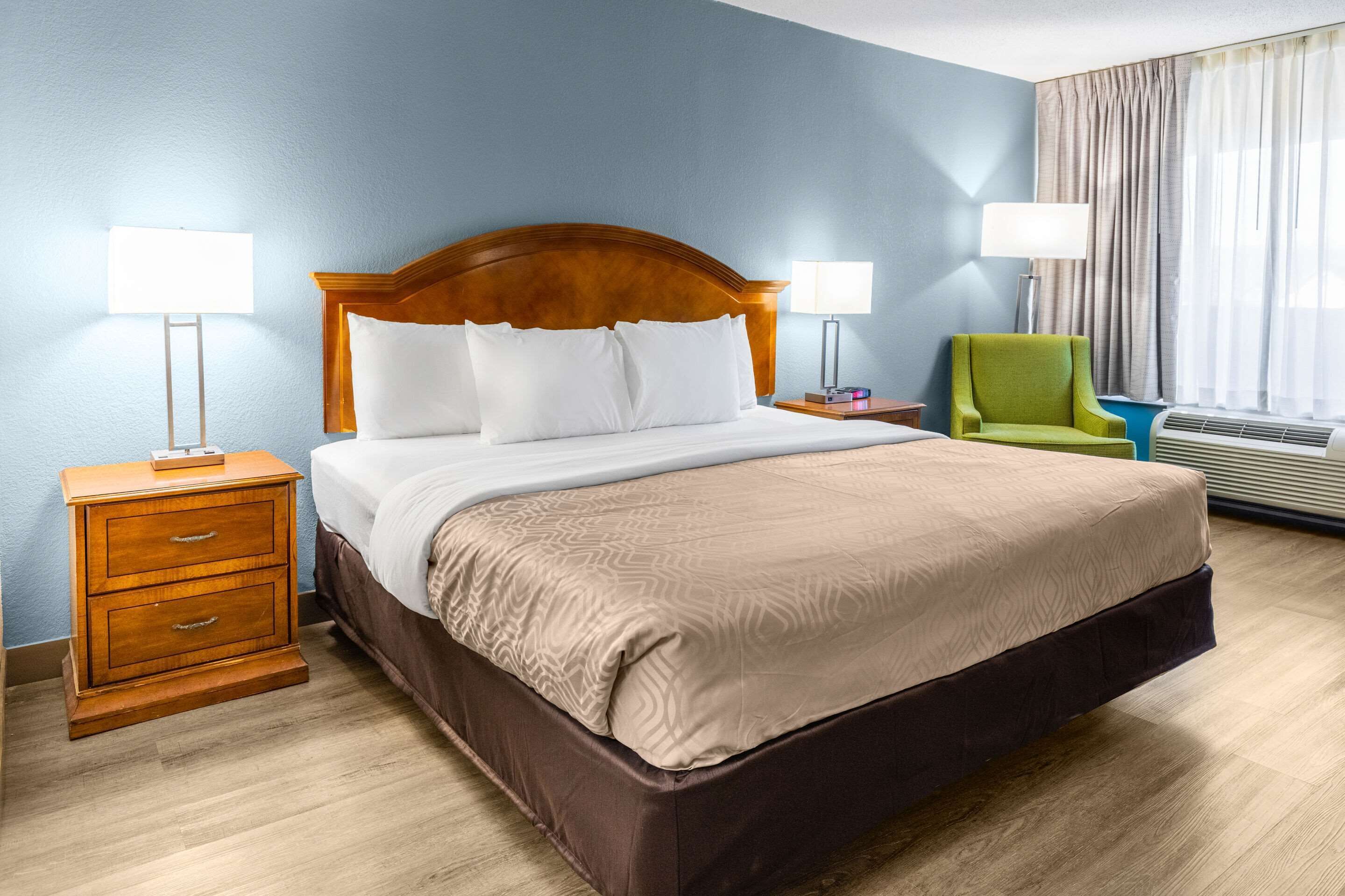 Une chambre d’hôtel avec un grand lit, une tête de lit en bois, deux tables de chevet avec des lampes, un fauteuil vert et une fenêtre avec des rideaux.