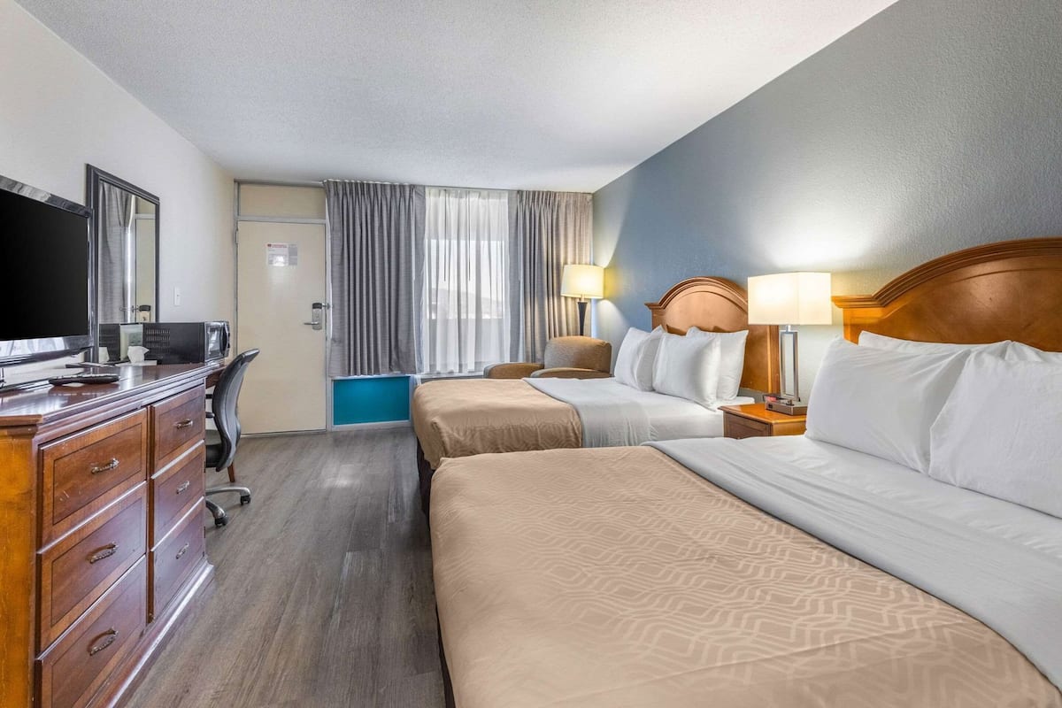 Ein Hotelzimmer mit zwei Betten, einem Schreibtisch mit Fernseher, einem Schminktisch und einem Fenster mit Vorhängen.