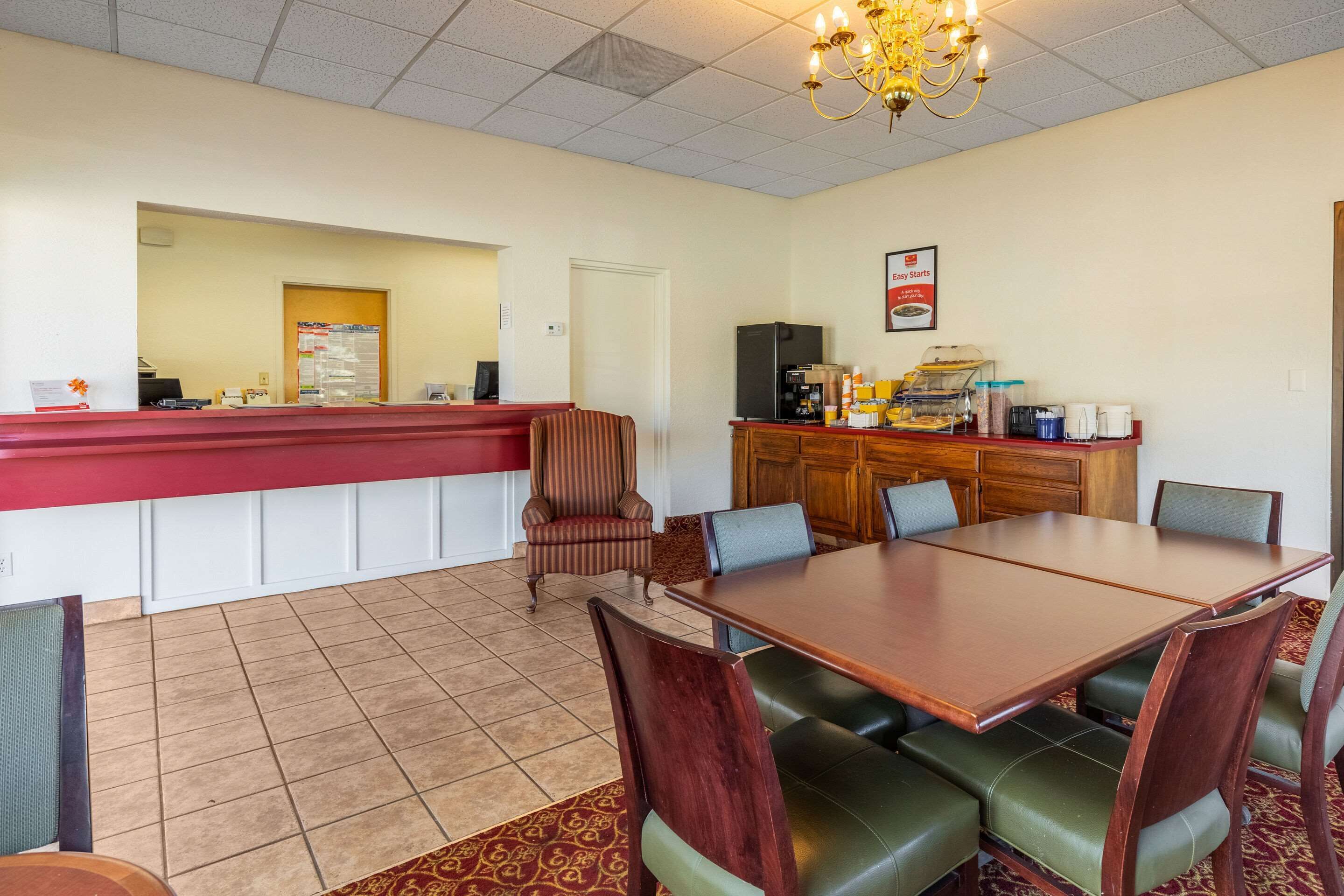 A hotel dining area with a long wooden table, green cushioned chairs, a red counter, and a kitchen area in the background.