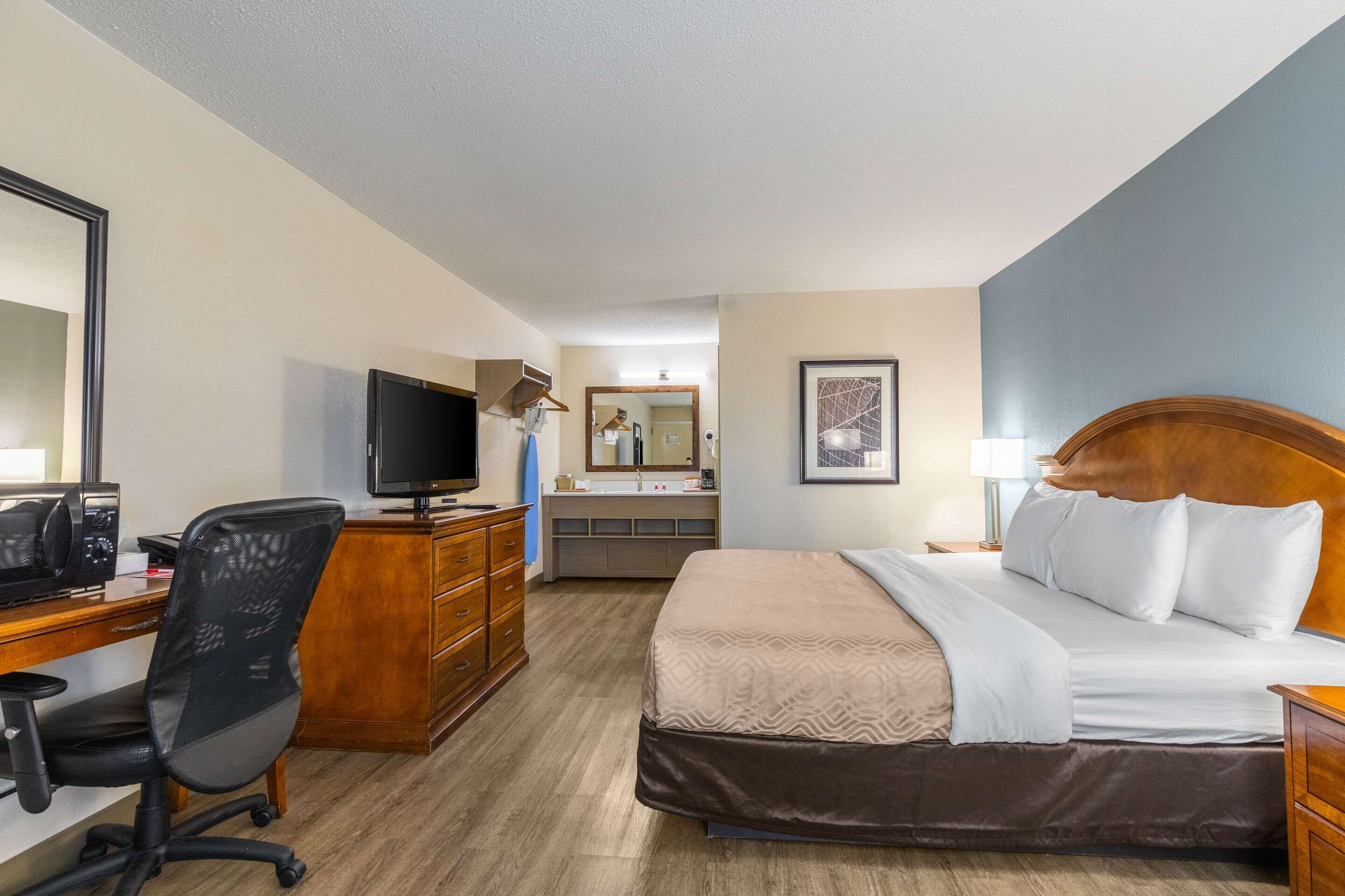 A hotel room with a bed, desk, chair, television, dresser, and a bathroom visible in the background.