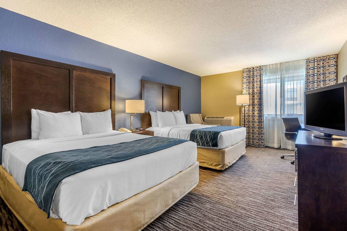 A hotel room with two beds, a desk, a TV, and a window with patterned curtains.