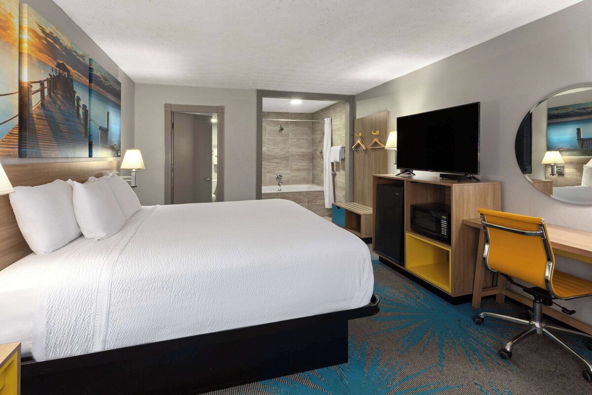 A hotel room with a large bed, a desk with a chair, a TV, and a shower area.