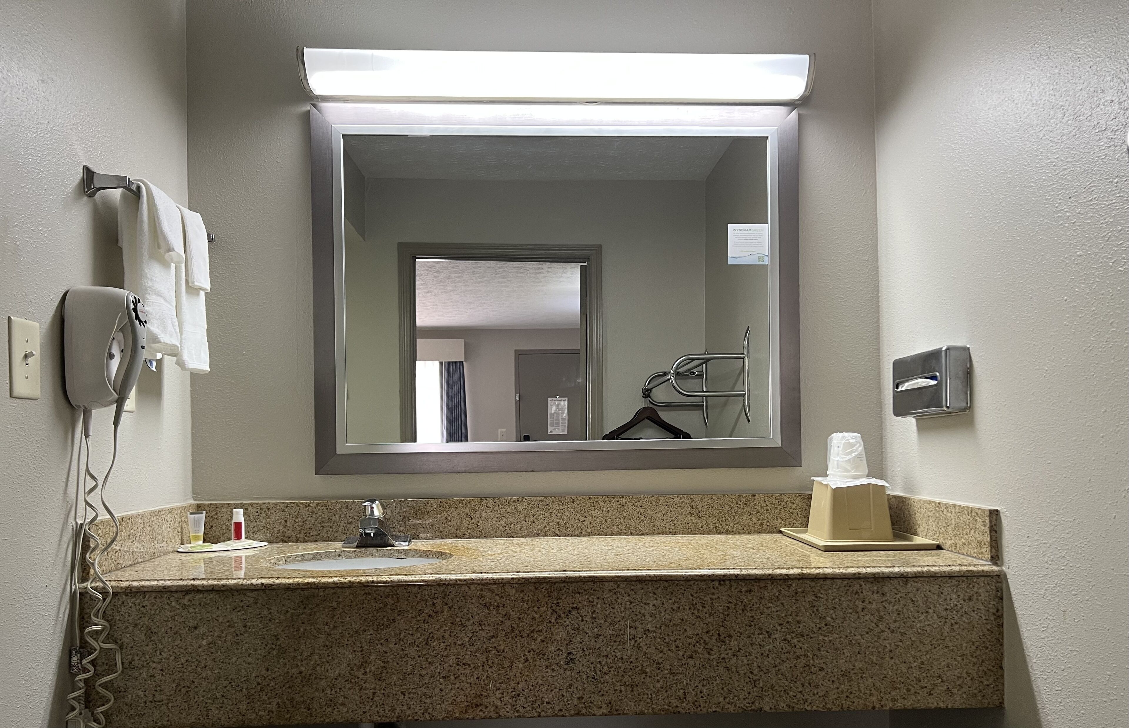 Une salle de bain dotée d’un comptoir en granit, d’un lavabo, d’un miroir, d’un sèche-cheveux, d’un porte-serviettes et d’un distributeur de mouchoirs.