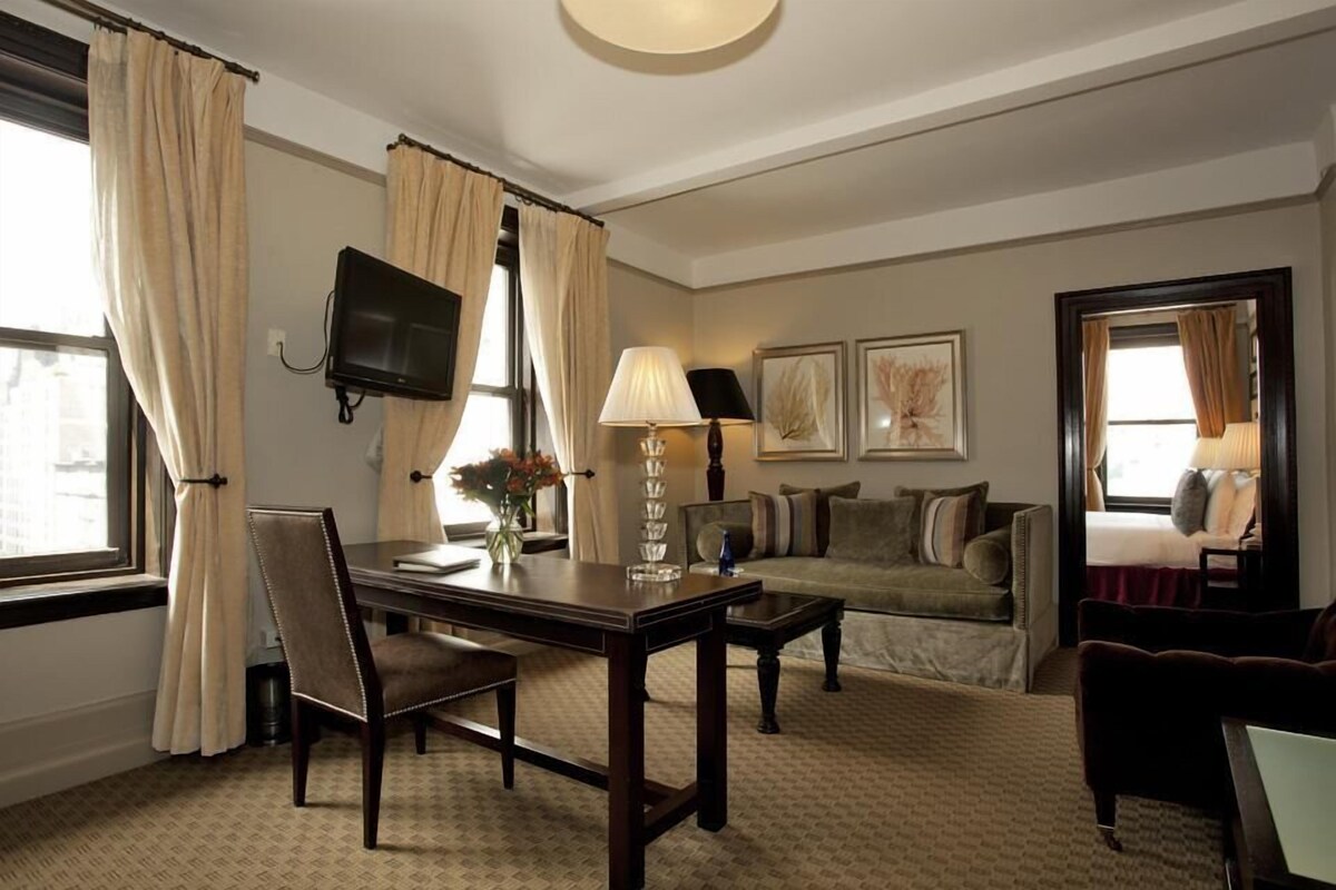 A hotel room with a wooden desk, a sofa, a TV mounted on the wall, and a bed with a window view.